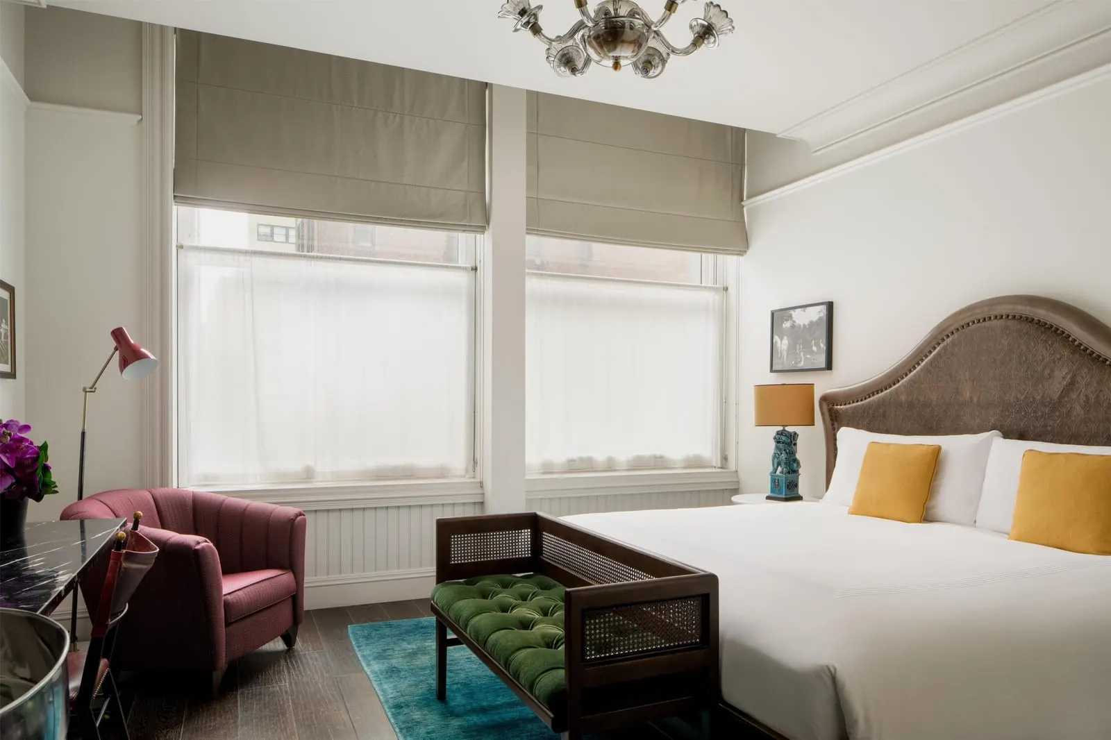 Premium King at The Beekman: a king bed with a leather headboard beside two tall windows with grey blinds, a green tufted bench at the foot of the bed and a burgundy armchair