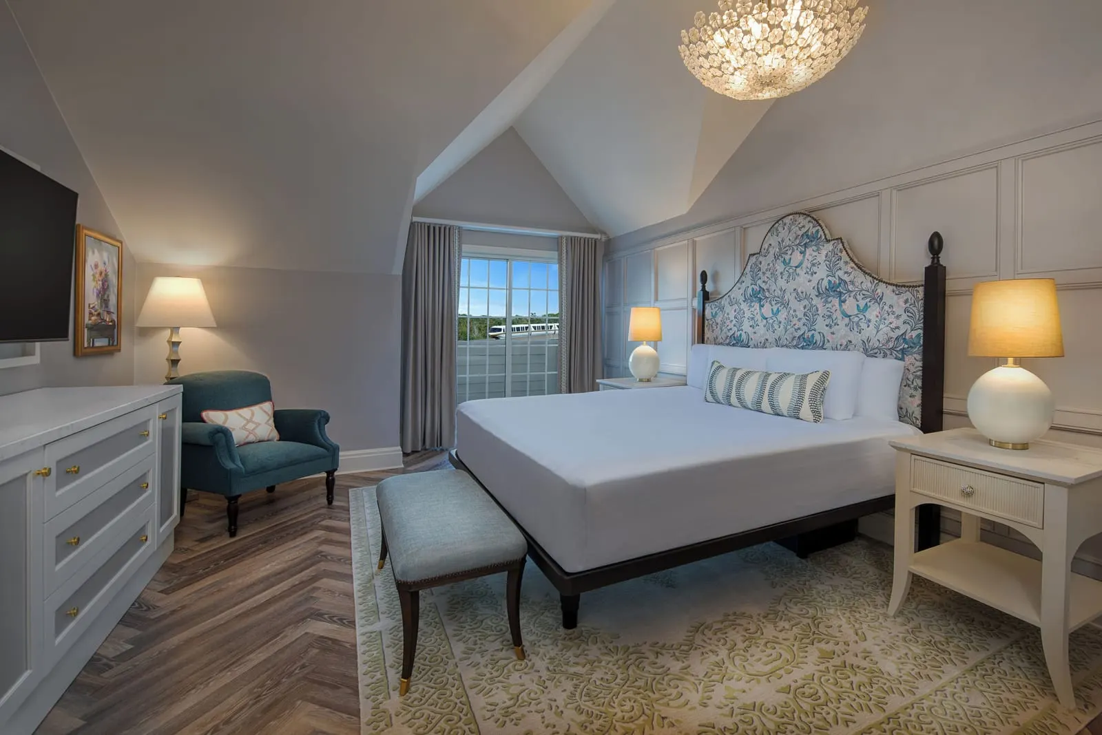 2 BR - Theme Park View - Club Level Access suite at Disney's Grand Floridian Resort & Spa: a king bed with a blue and white patterned headboard under a sloped ceiling, with a padded bench, a teal armchair and a white chest of drawers