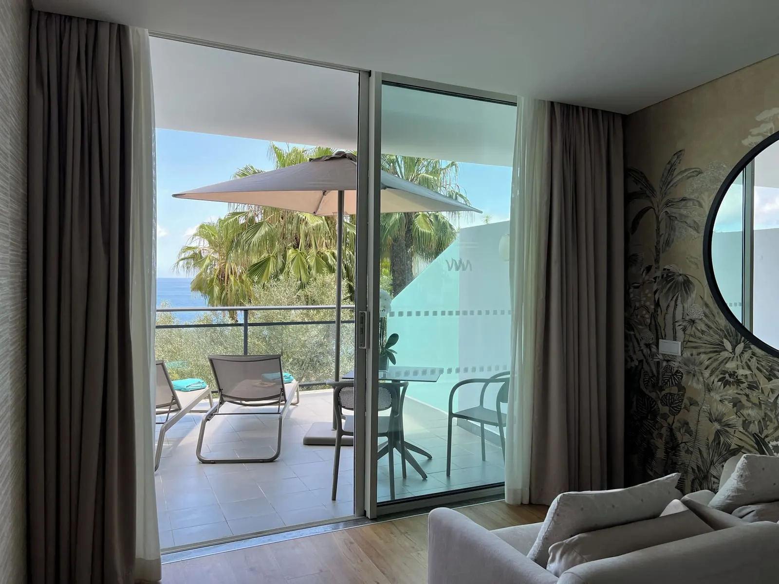 VidaMar Resort Hotel Madeira Terrace Junior Suite Sea View：ヤシの木と海を望む、ラウンジャー2脚、パラソル、テーブルを置いたパティオへ続くガラス扉