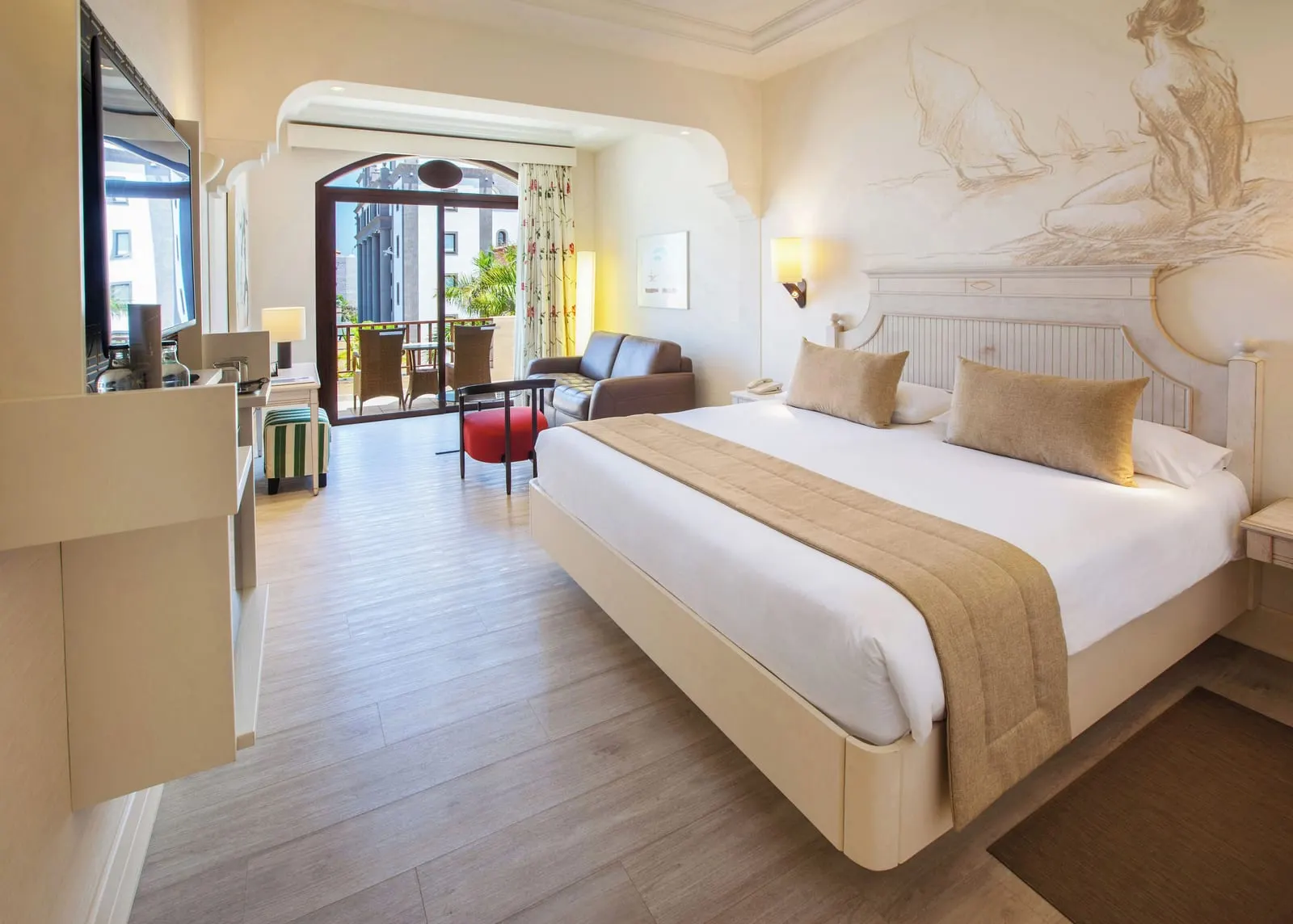 Double Standard at Lopesan Villa del Conde Resort & Thalasso: a king bed under a sketched mural, a small leather sofa, a red chair and an arched glass door to a balcony with wicker chairs