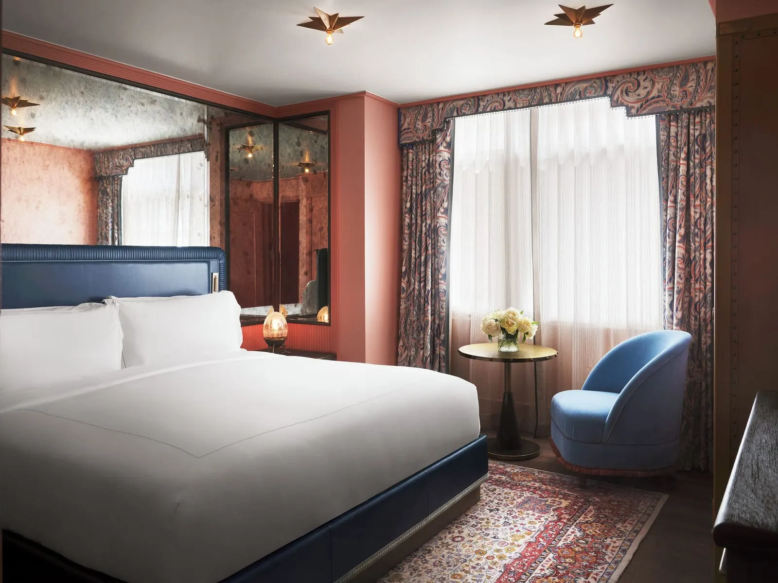 Parlour Suite at The Fifth Avenue Hotel: a bedroom with a bed in white linen and a dark blue headboard, a mirrored wall, a blue armchair and a round table in front of tall windows with paisley curtains