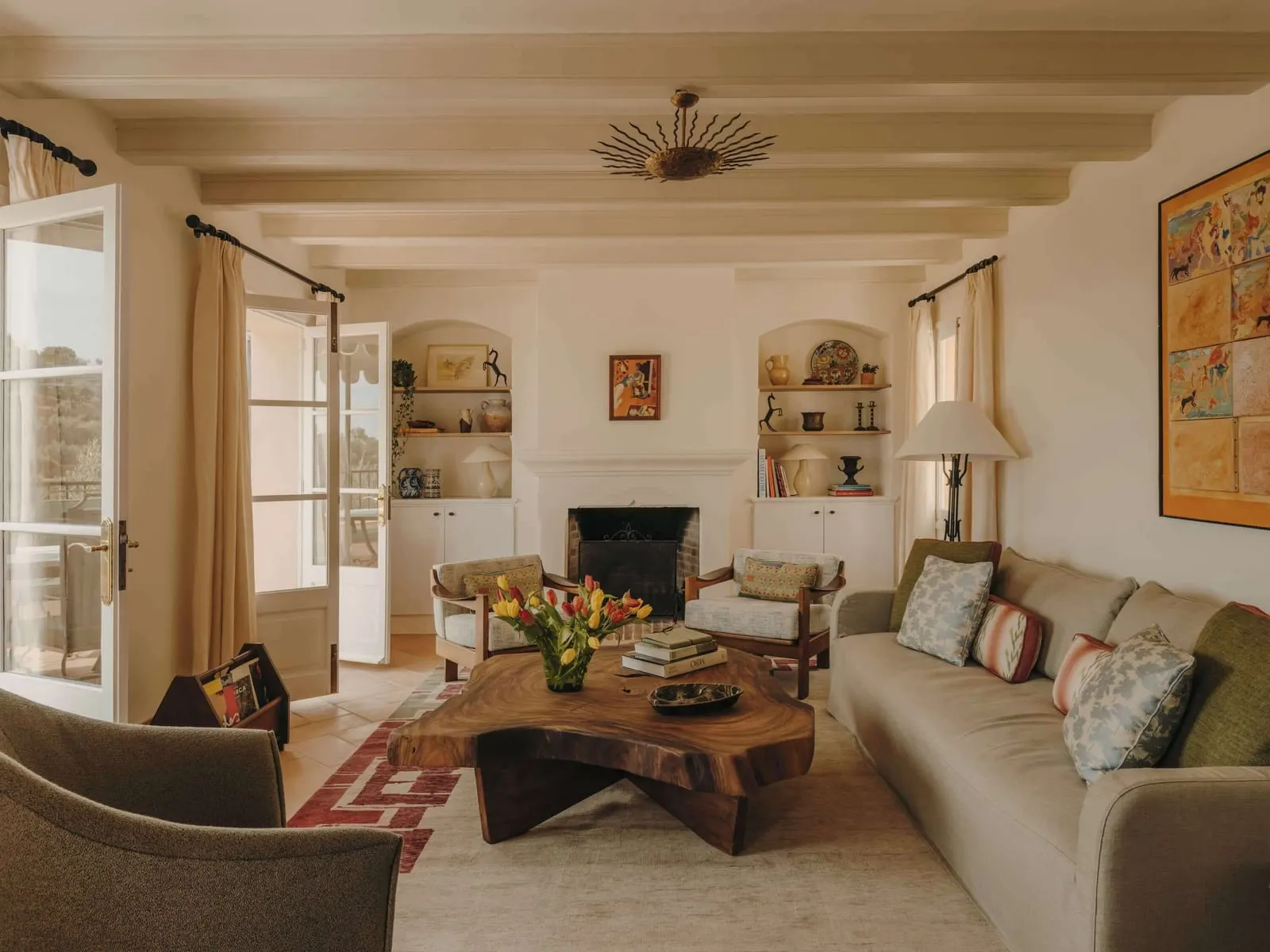Villa Robert Graves at Belmond La Residencia: a living room with a fireplace, a sofa, two armchairs and a slab wooden coffee table under painted beams, with French windows to a balcony