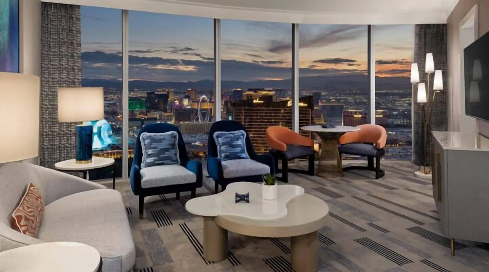 Noble Suite Strip Great View at Fontainebleau Las Vegas: the living room, with a curved sofa, two navy armchairs and a round table with two chairs by a wall of windows onto the Strip at dusk