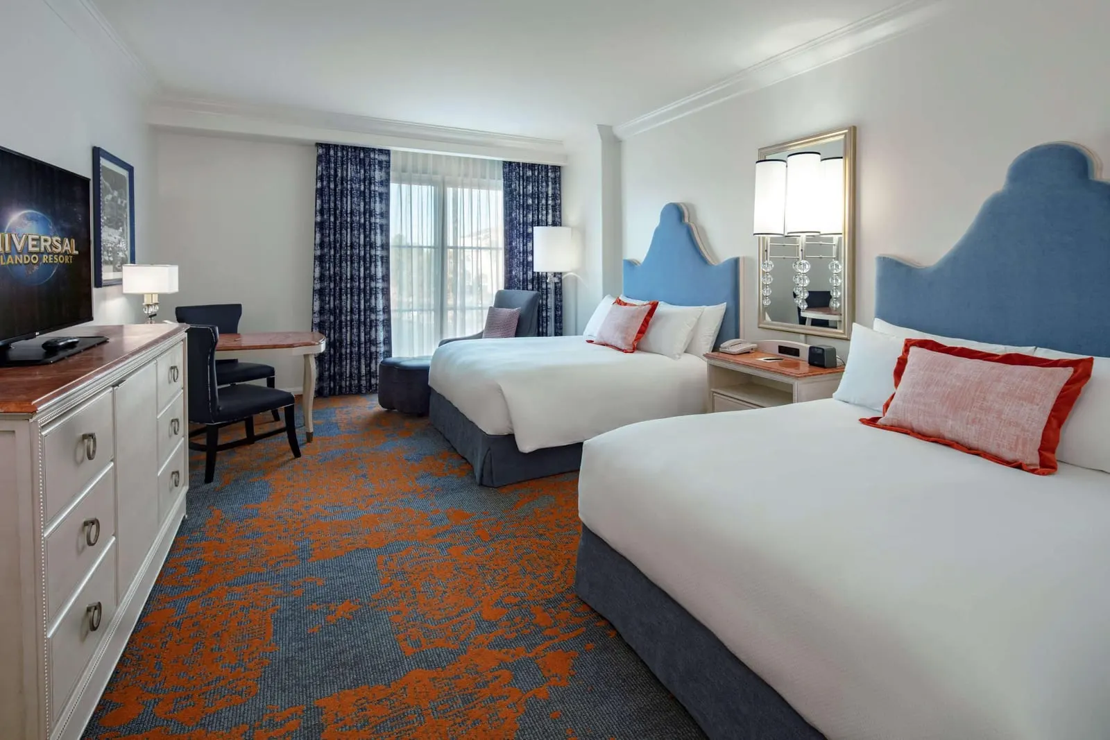 Garden View 2 Queen Room at Loews Portofino Bay Hotel: two queen beds with blue upholstered headboards, a dresser with a television, a writing desk and an armchair beside a window with the sheer curtains drawn