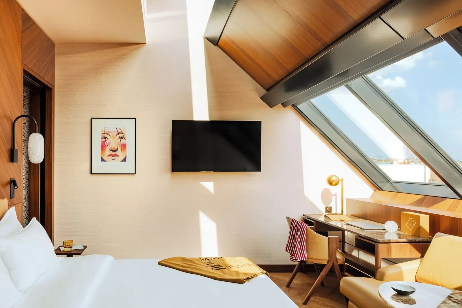 Deluxe Atelier at Hôtel Madame Rêve: a king bed, a wall-mounted television, a writing desk and a leather armchair beside a tall sloping atelier window framed in timber