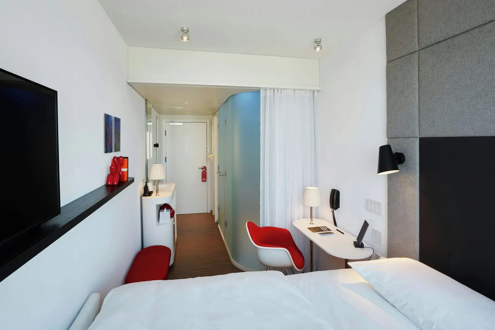 Guest room, 1 King, High floor at citizenM New York Bowery: a king bed with white linen against a grey padded headboard, a red armchair and small desk, a TV on a black shelf and a frosted glass shower pod leading to the door