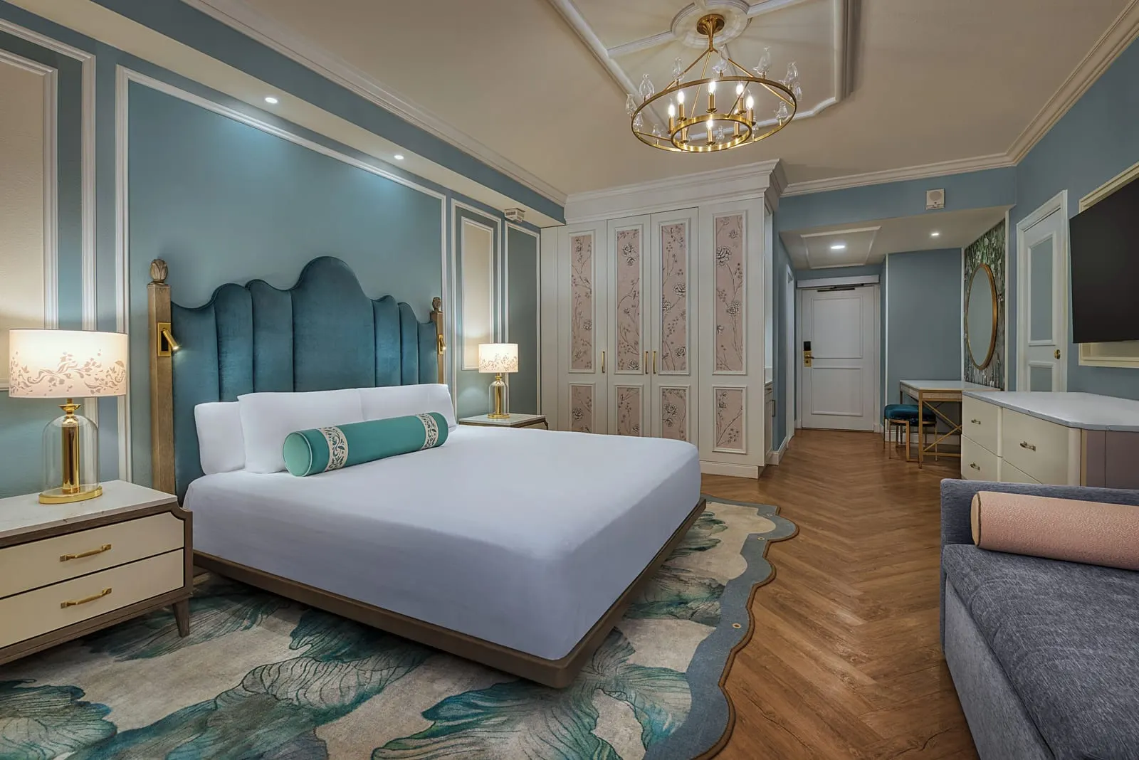 Deluxe King Room - Club Level at Disney's Grand Floridian Resort & Spa: a king bed with a scalloped teal velvet headboard under a chandelier, a wardrobe with painted floral panels, a desk and a blue sofa