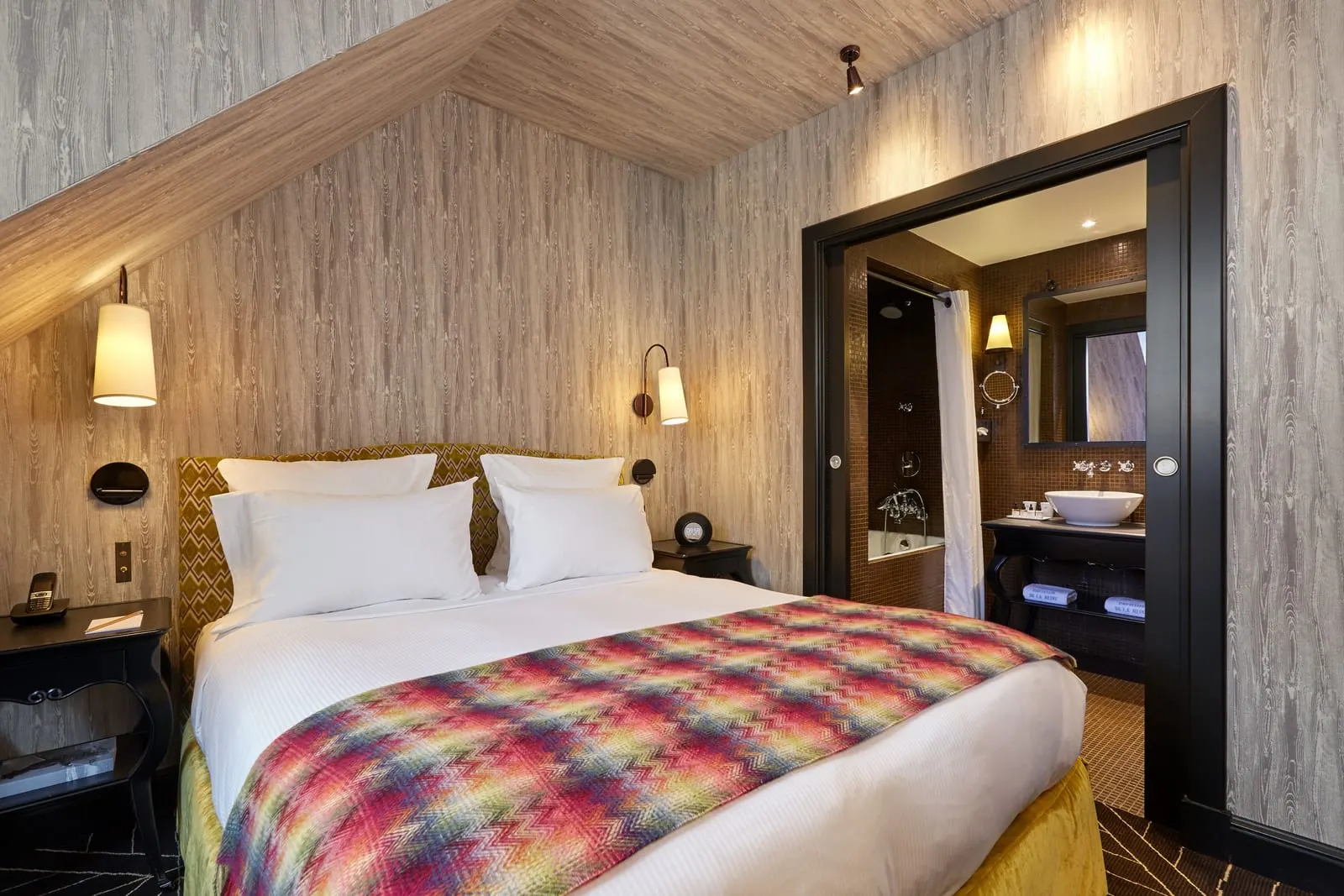 Classic Room at Le Pavillon de la Reine: a double bed with a multicoloured zigzag throw under a sloping wood-effect ceiling, and the bathroom through an open sliding door