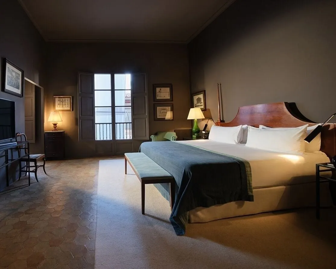 Junior Suite im Can Cera: ein großes Bett mit einer blaugrauen Tagesdecke und einer Bank, ein hohes Fenster mit Fensterläden und Balkongeländer und ein Boden mit sechseckigen Fliesen