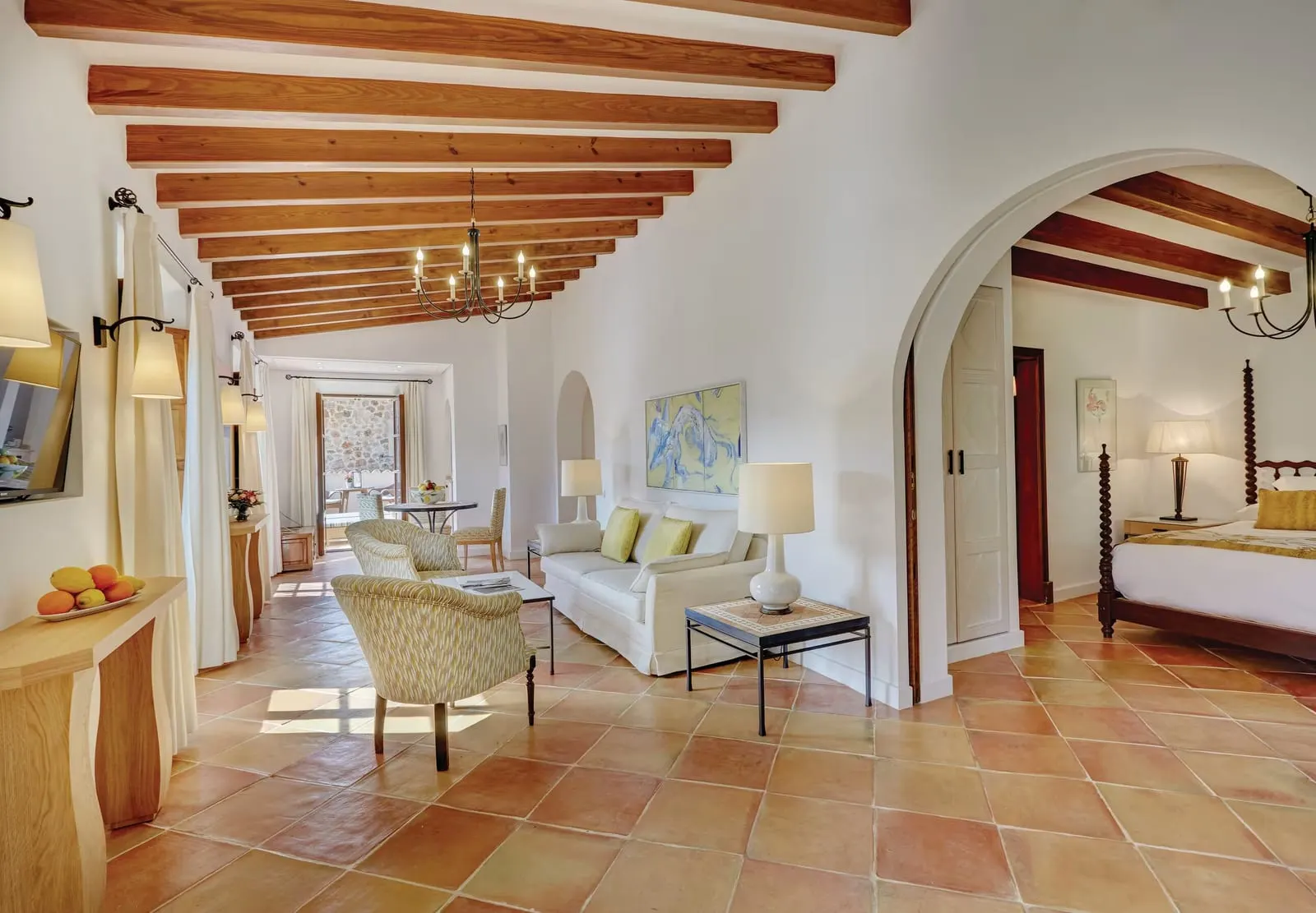 Deluxe Suite at Belmond La Residencia: a living room with a cream sofa, patterned armchairs and a chandelier on a terracotta tiled floor, with an arch leading to a four-poster bedroom