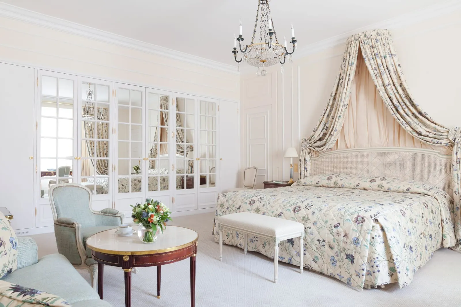 Suite Princière at Le Bristol Paris: a bed under a draped canopy with a floral quilted cover, a round table with armchairs and a wall of mirrored wardrobe doors
