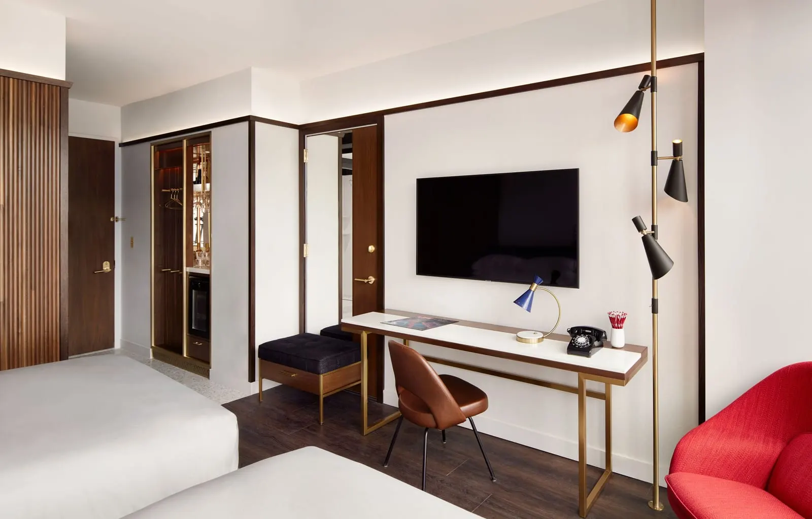 Standard Double Queen at TWA Hotel: two queen beds beside a long desk with a rotary phone, a wall-mounted television and a brown leather chair, with a red armchair at the edge of the frame