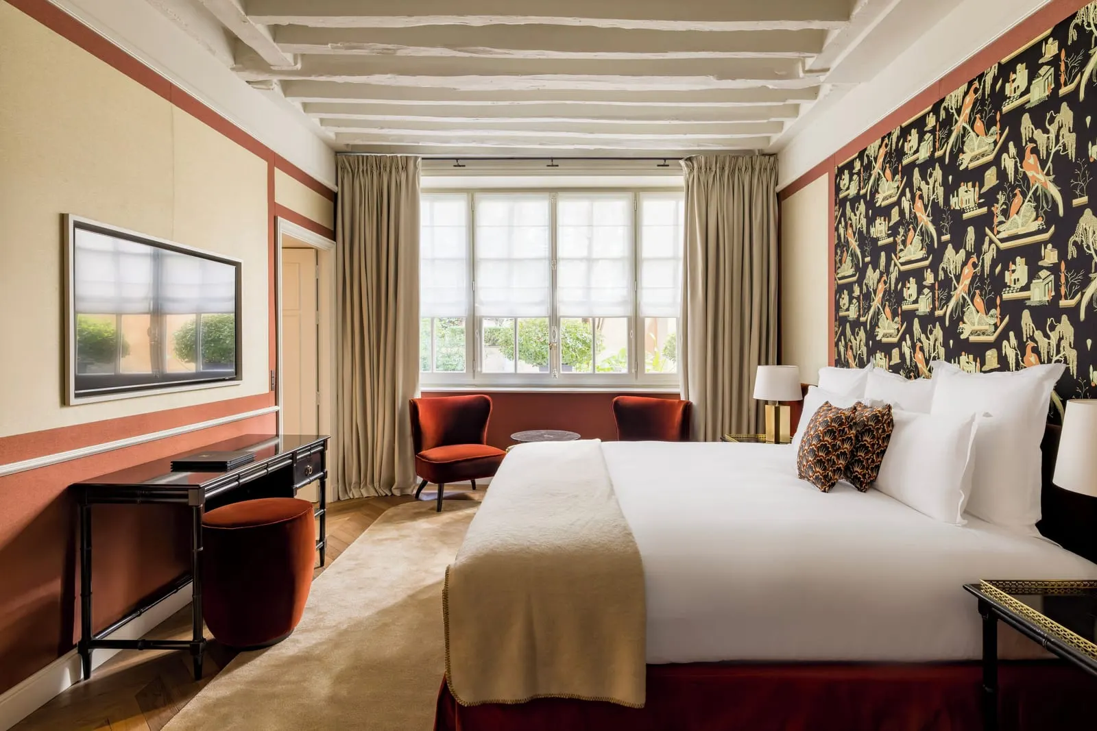 Suite at Le Pavillon de la Reine: the bedroom, with a double bed against a bird-print wall panel, white ceiling beams, two velvet armchairs by a wide window and a writing desk
