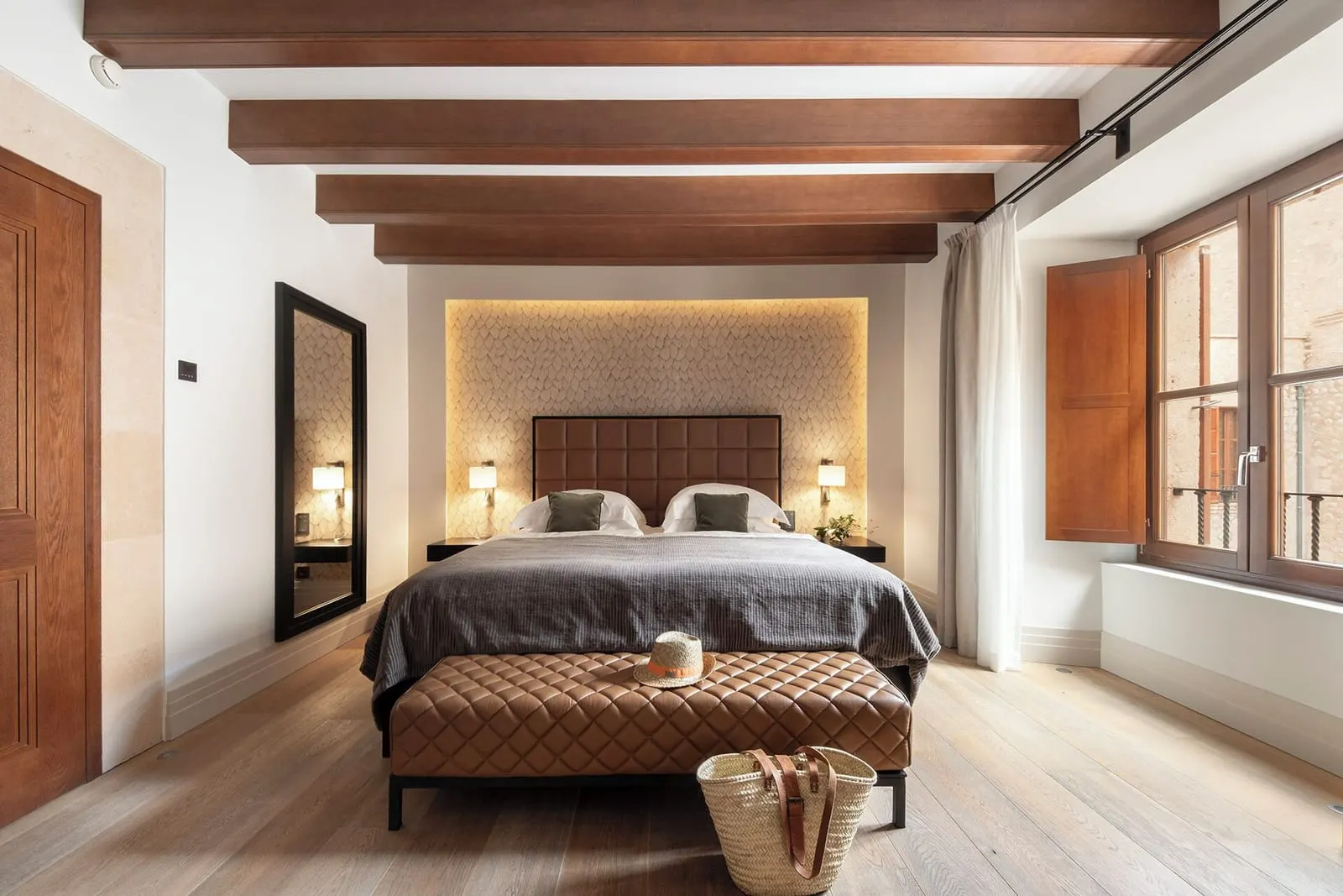 Estate Courtyard im Castell Son Claret: ein Bett mit einer gesteppten Lederbank am Fußende unter dunklen Holzbalken, mit einem beleuchteten Paneel hinter dem Kopfteil und einem Fenster mit Fensterläden und Steinmauerwerk dahinter