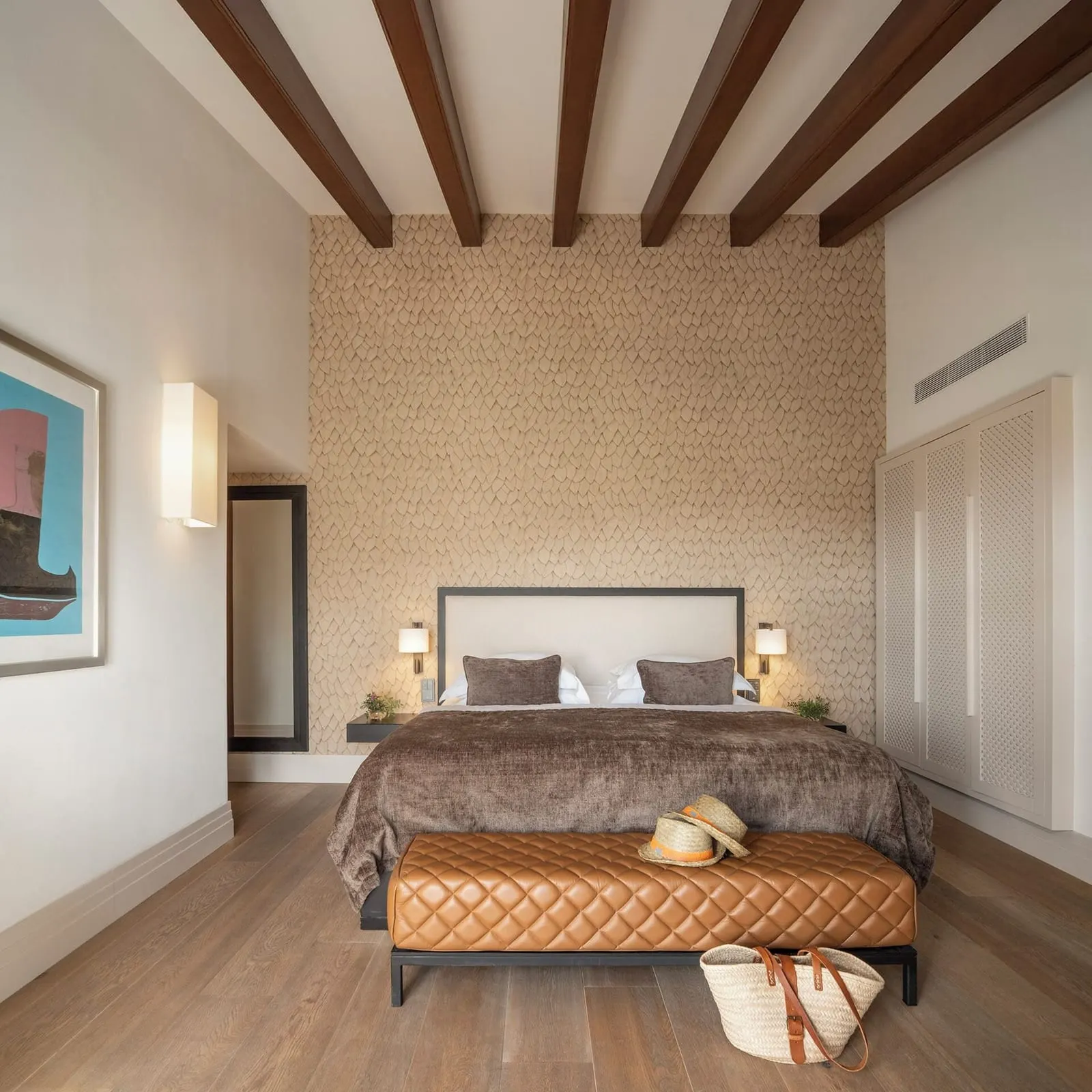 Deluxe Double at Castell Son Claret: a bed with a quilted leather bench under exposed wooden beams, a textured wall behind the headboard and a lattice-fronted wardrobe