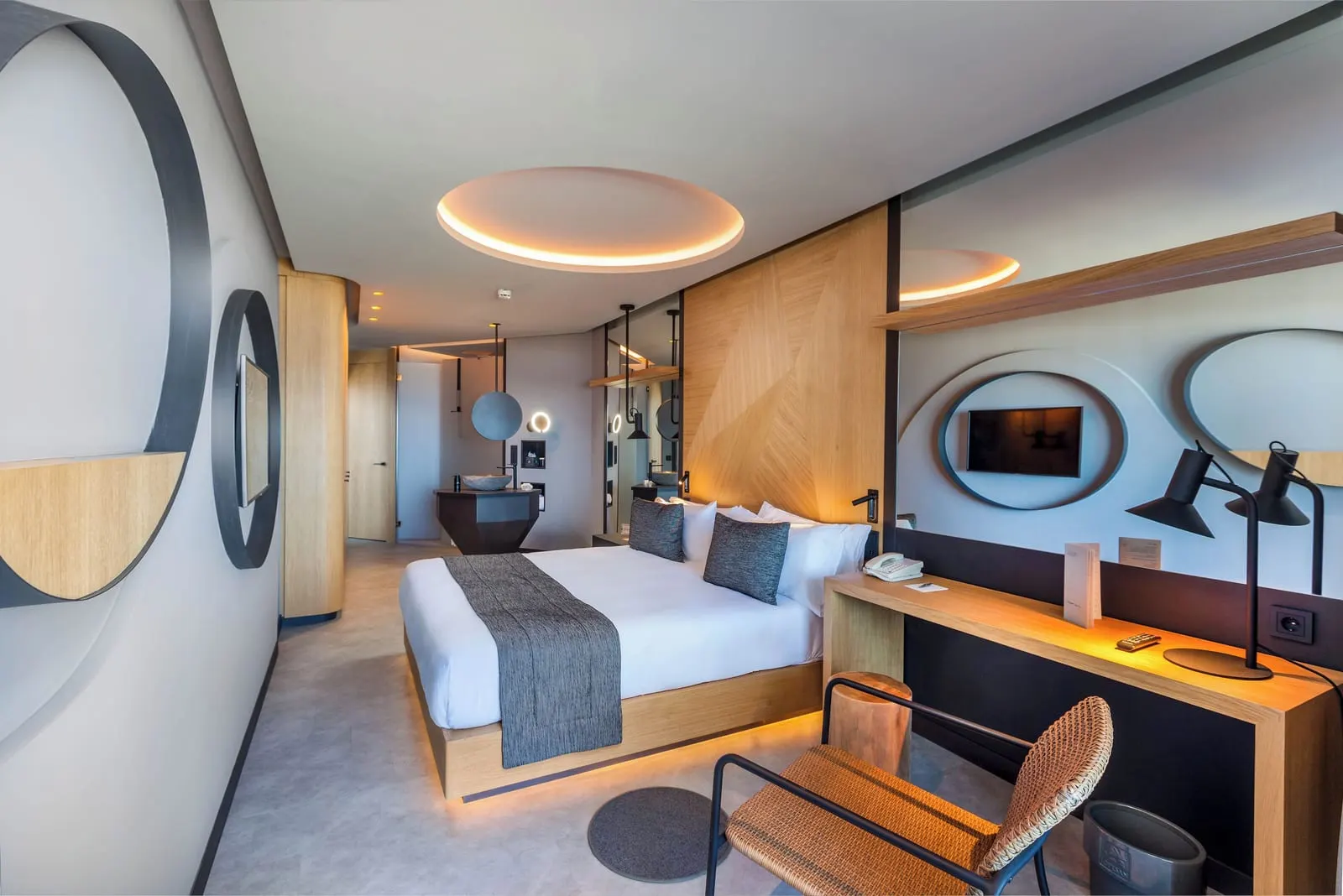 Double Deluxe at Hotel Faro, a Lopesan Collection Hotel: a double bed with a grey throw, a long wooden desk with two black lamps, and a stone washbasin open to the room by the entrance