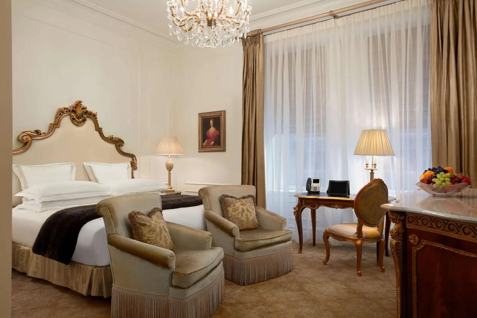 Plaza King at The Plaza: a king bed with a gilded headboard, two velvet armchairs, a writing desk and a fruit bowl on a marble topped chest, under a crystal chandelier
