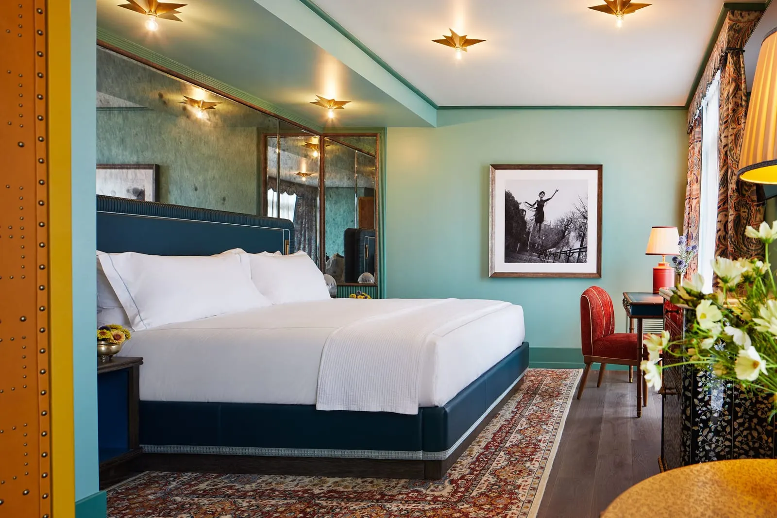 The Grand King at The Fifth Avenue Hotel: a king bed with white linen and a dark blue headboard under a mirrored wall, a framed black and white photograph, and a desk with a red chair by the window