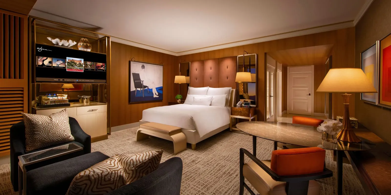Wynn Panoramic View King at Wynn Las Vegas: a king bed with a tall padded headboard and a bench at its foot, two dark armchairs, an oval table with orange-backed chairs and a cabinet holding the television