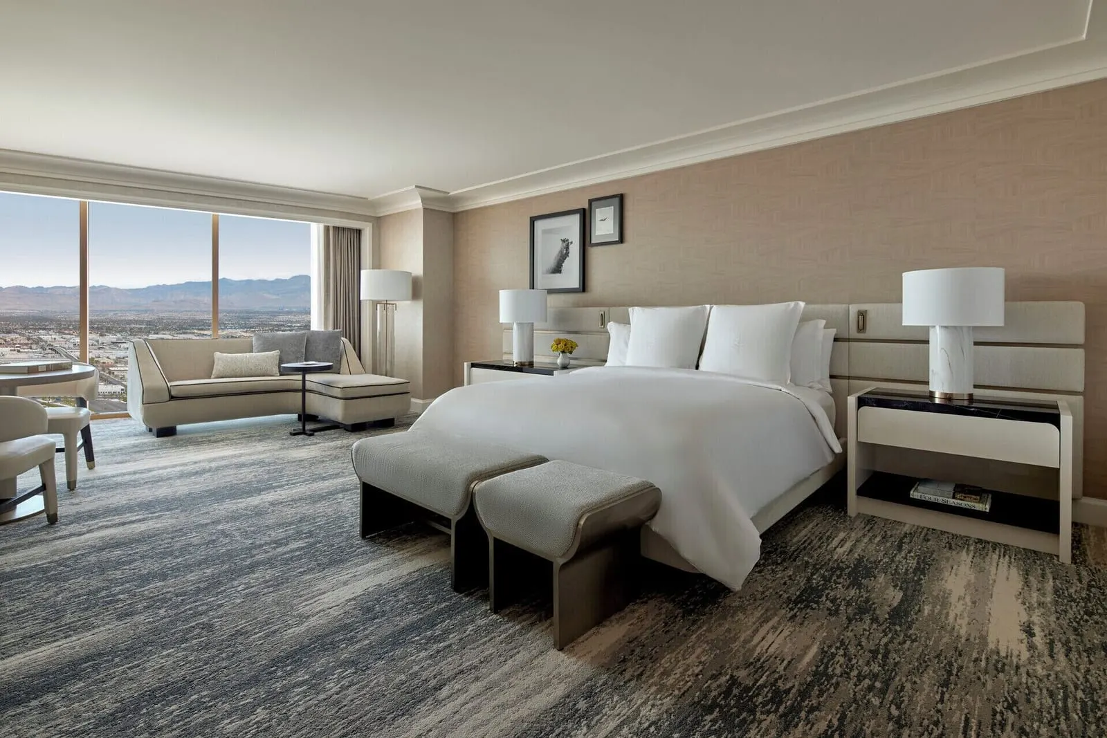 Stadium View Room at Four Seasons Hotel Las Vegas: a king bed with a two-part bench at its foot, and a curved chaise sofa and a round table by a floor-to-ceiling window