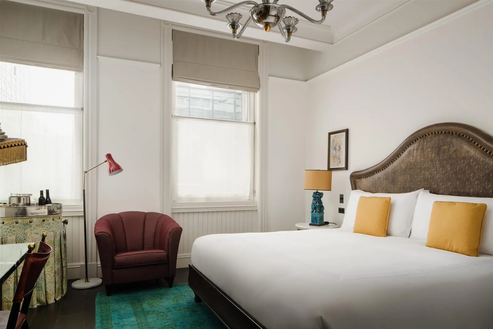 Deluxe King at The Beekman: a king bed with a studded leather headboard beside two tall windows, a burgundy armchair, a red reading lamp and a chandelier