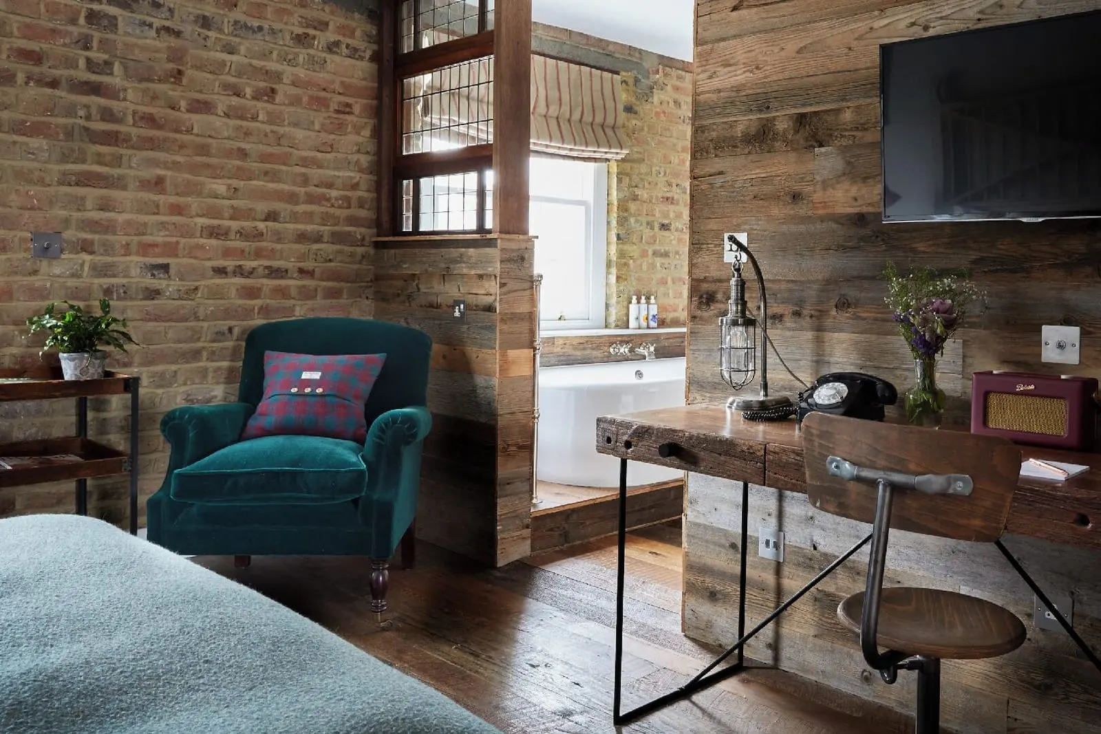 The Loft at Artist Residence London: a teal velvet armchair, a desk and the bath behind a timber partition