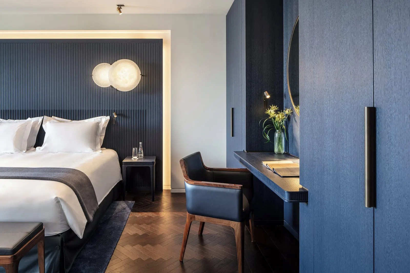 Eiffel View Deluxe Balcony Room at Mandarin Oriental Lutetia, Paris: a double bed against a dark blue fluted headboard with two round glass wall lights, a leather desk chair at a slim writing desk and dark blue built-in wardrobes on a herringbone wood floor