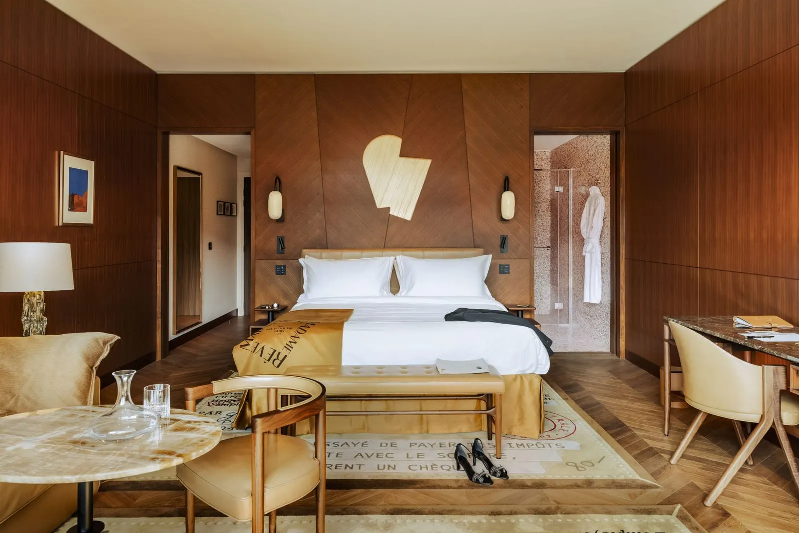 Junior Suite Family at Hôtel Madame Rêve: a king bed with a leather bench at its foot against walnut panelling, a round stone-topped table and armchair, a writing desk, and doorways to a mirrored hall and a mosaic-tiled shower room