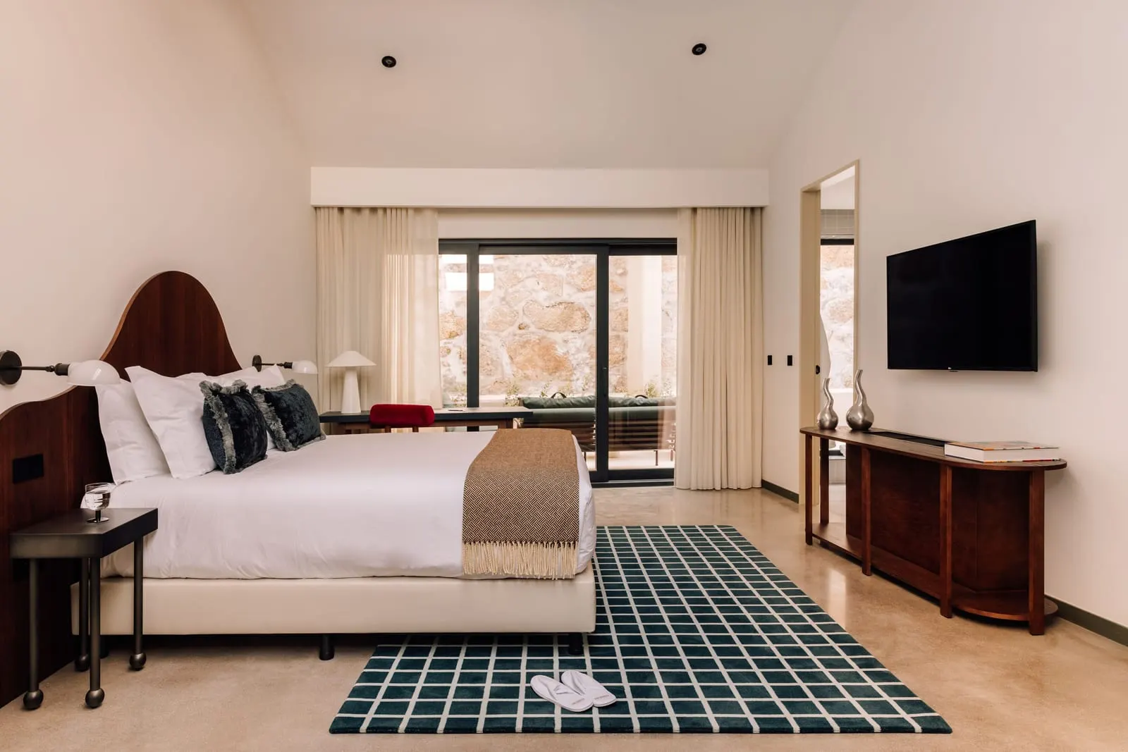 One Bedroom River Suite at The Rebello: the bed with a curved wooden headboard, a sideboard and glass doors at the far end