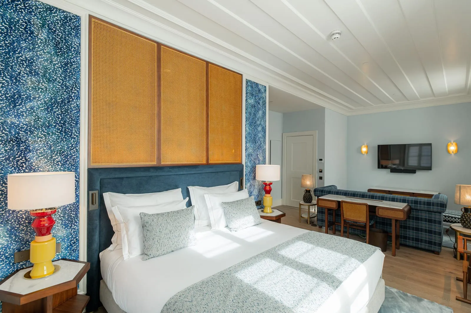 Junior Suite at Palácio Ludovice: the bed below rattan panels and blue patterned walls, with a desk and a tartan sofa beyond