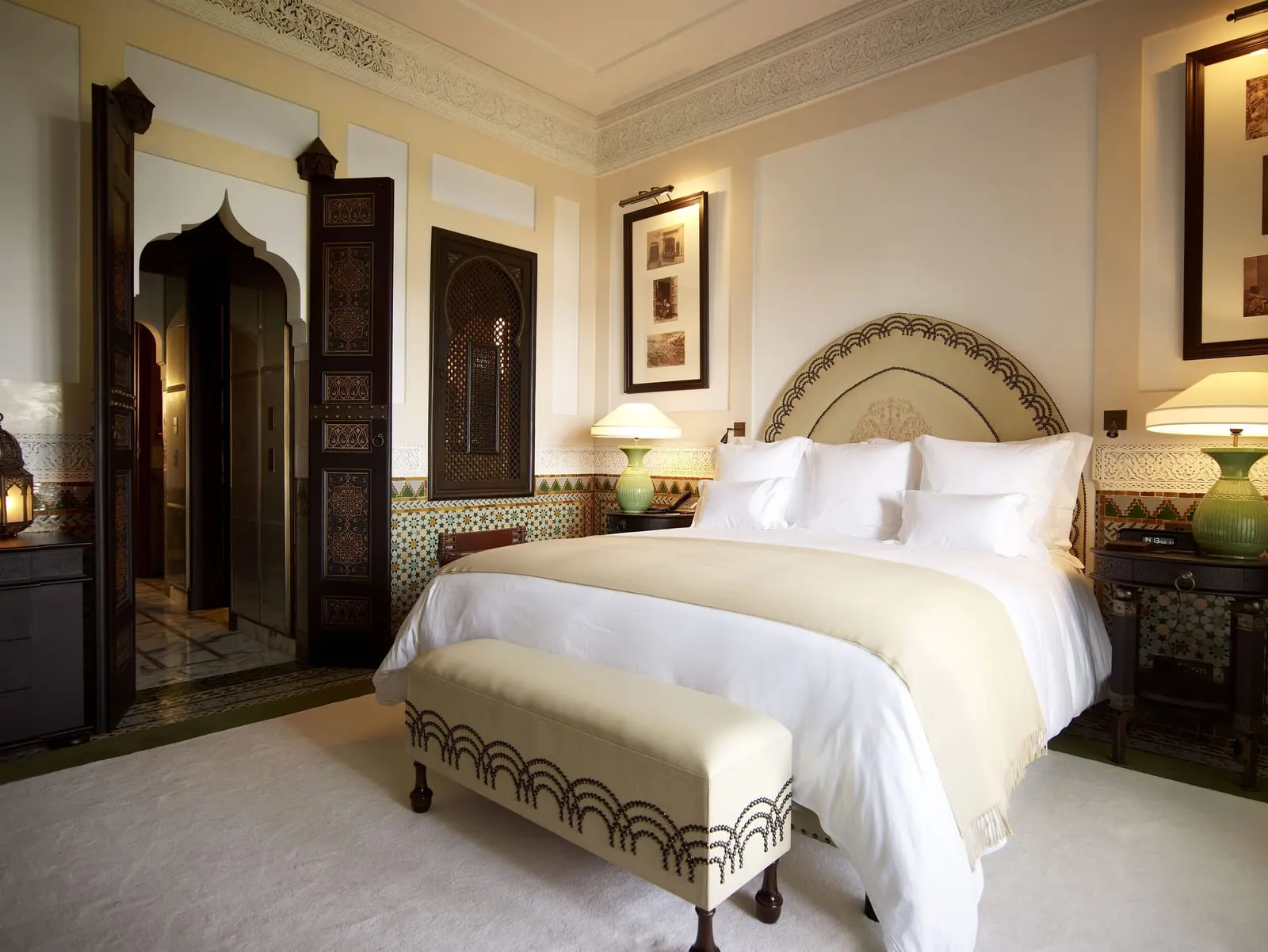 Koutoubia Deluxe Room at La Mamounia: a king bed under an arched carved headboard, with a cream bench, green lamps and zellige tiles
