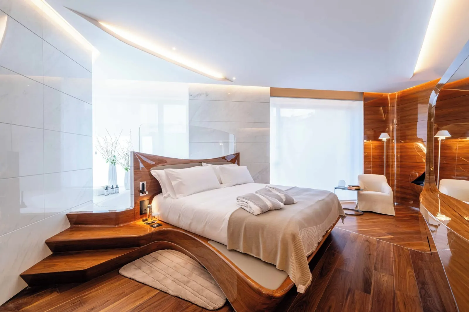 Deluxe room at ROMEO Roma: a bed on a curved walnut platform with steps, marble walls and an armchair by the window