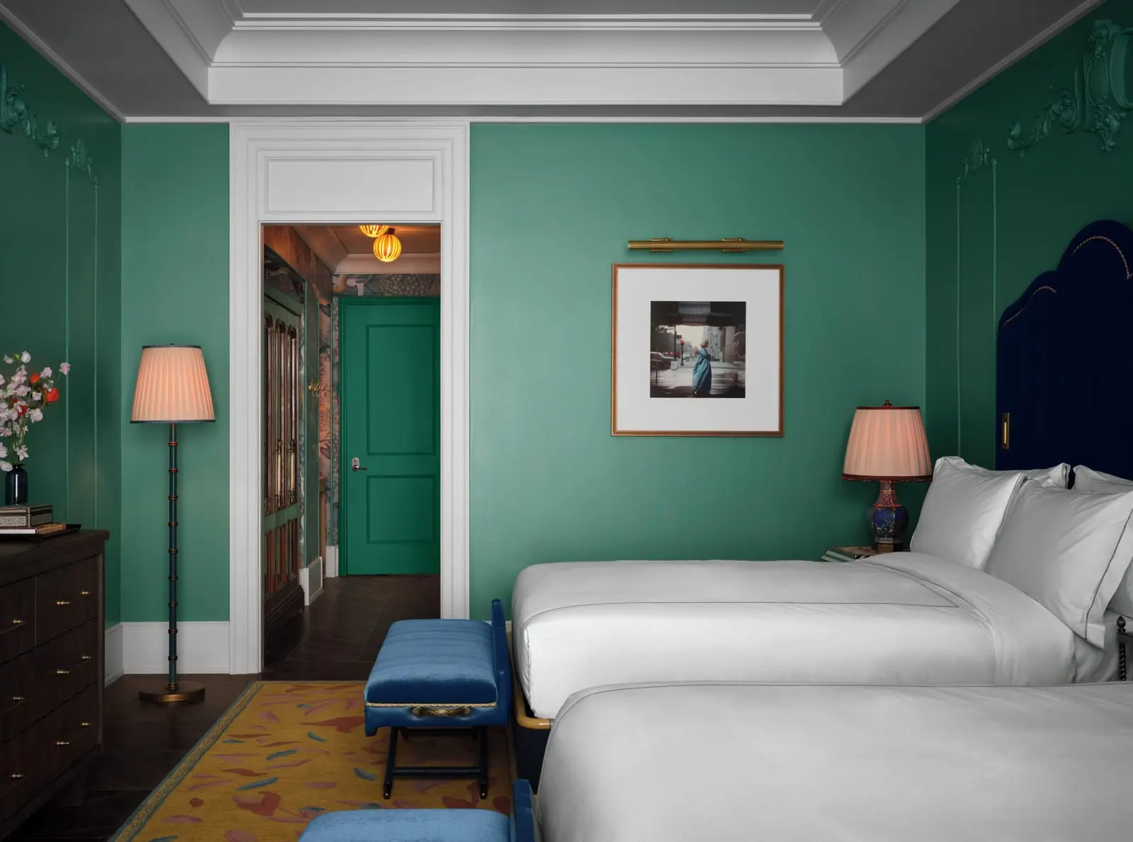 The Mansion Suite Double at The Fifth Avenue Hotel: two beds with white linen in a green room with plaster mouldings, a blue bench, a chest of drawers and a framed photograph, and a doorway to a hall behind