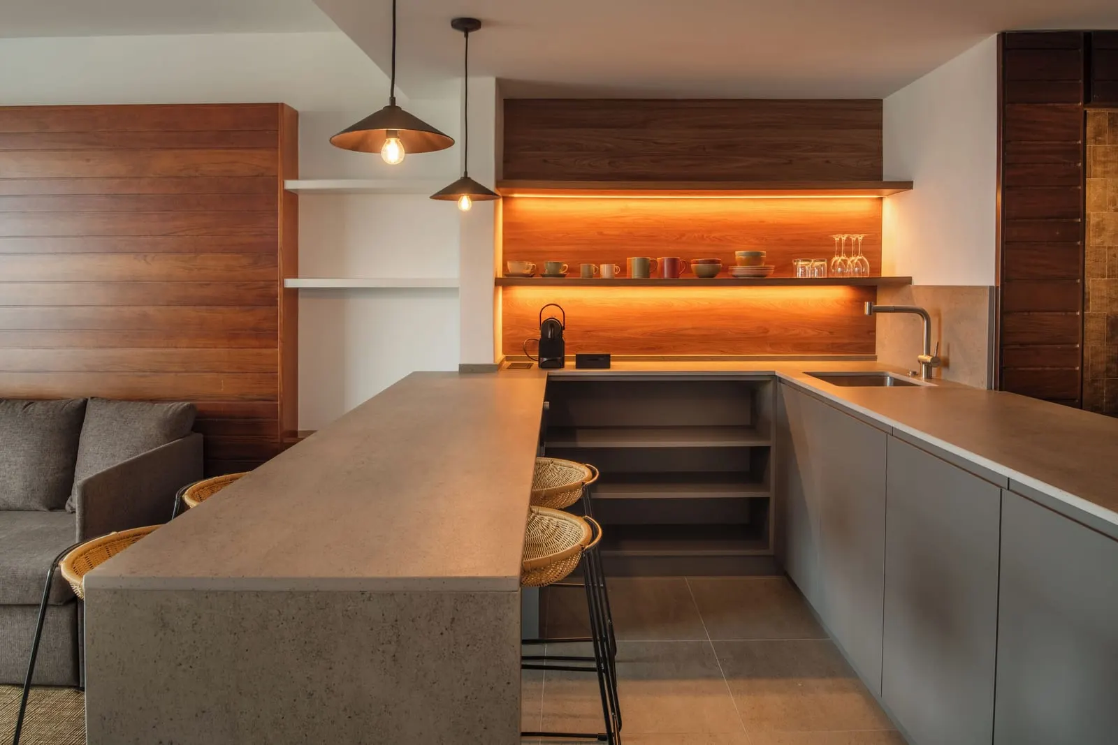 Junior Suite at Salobre Hotel Resort & Serenity: a small kitchen with a breakfast bar and rattan stools, a sink and lit shelves of cups and glasses, beside a grey sofa