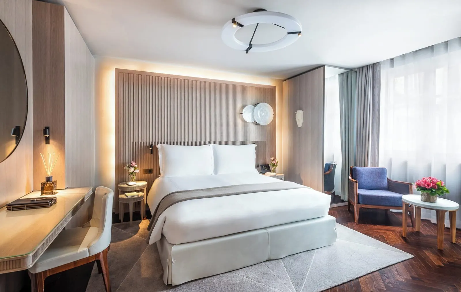 Grand Superior Room at Mandarin Oriental Lutetia, Paris: a double bed against a pale fluted oak headboard with two round glass wall lights, a writing desk, and a blue armchair and side table by tall windows with sheer curtains