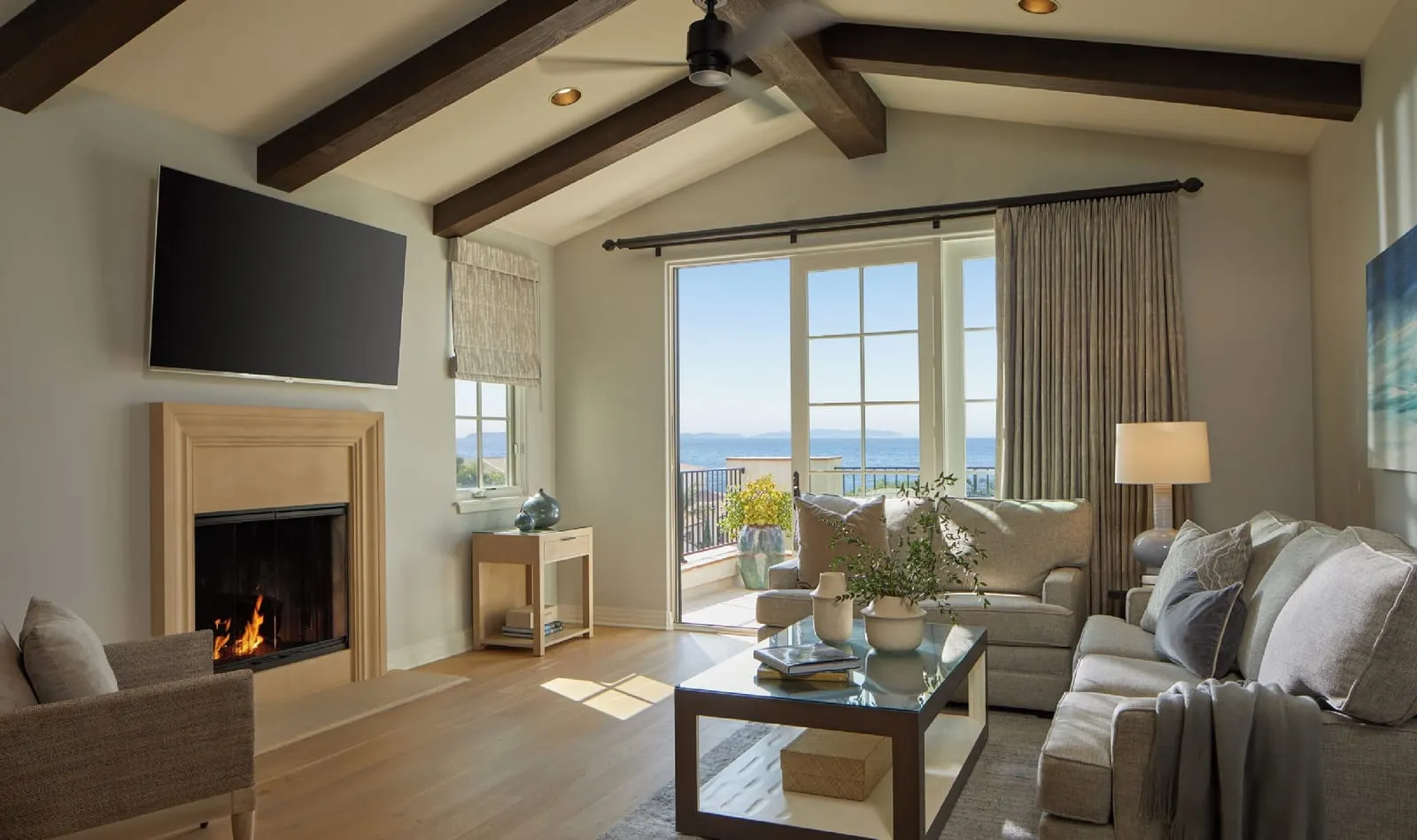 One-Bedroom Casita, Premier Ocean View at Terranea Resort: a living room with a fireplace, a wall-mounted television, grey sofas and glass doors onto a balcony, under dark ceiling beams