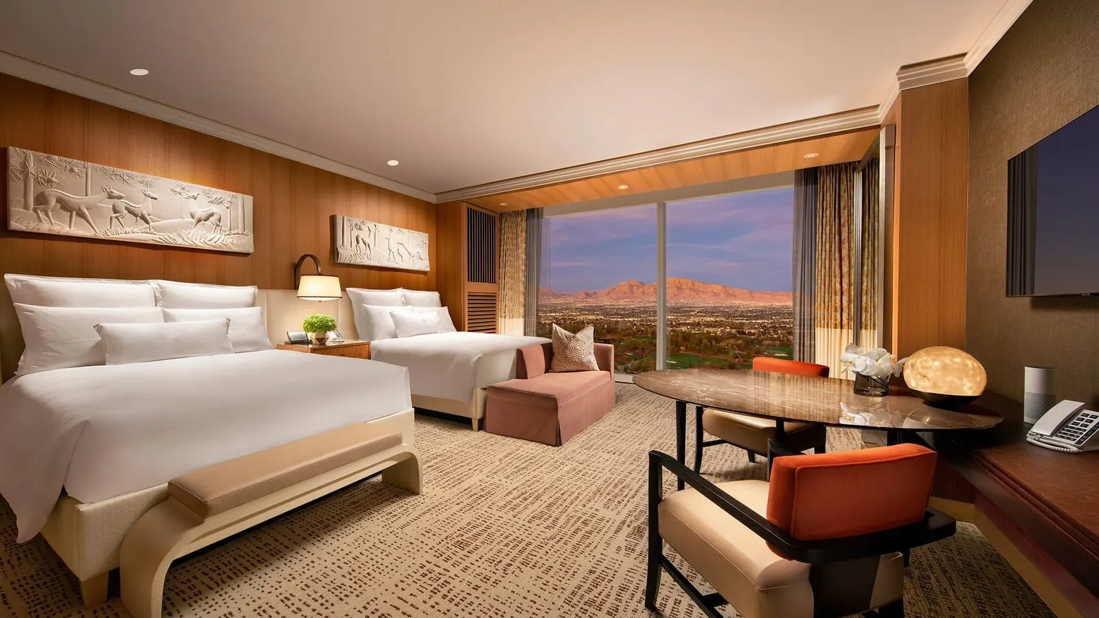 Wynn Panoramic View Two Queens at Wynn Las Vegas: two queen beds below carved relief panels, a small sofa and an oval table with two orange-backed chairs by a floor-to-ceiling window