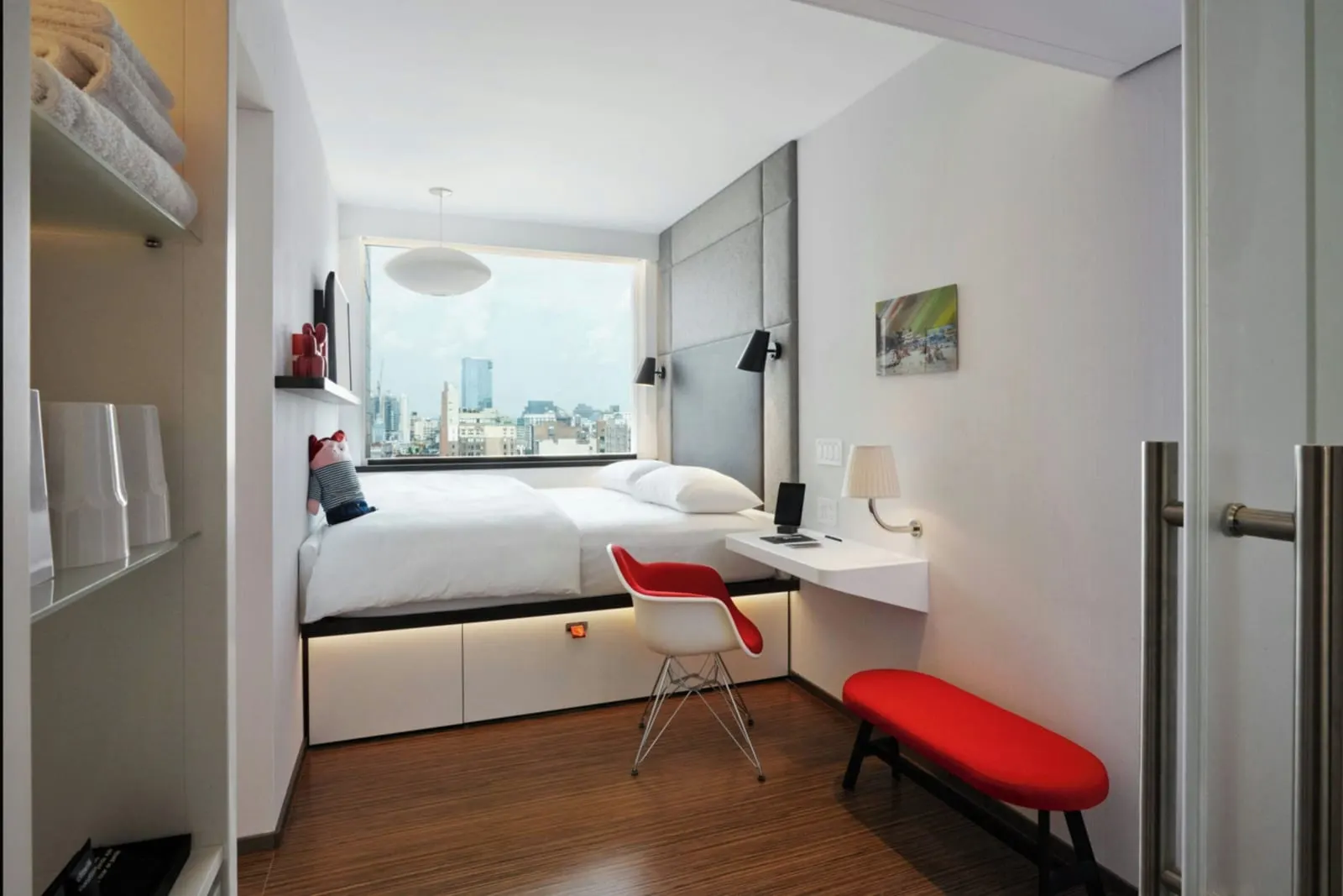 Guest room, 1 King, Premium view at citizenM New York Bowery: a king bed with white linen under a wide window with rooftops and a glass tower beyond, a red and white chair, a wall desk and a red bench in front, and towels on a shelf at left