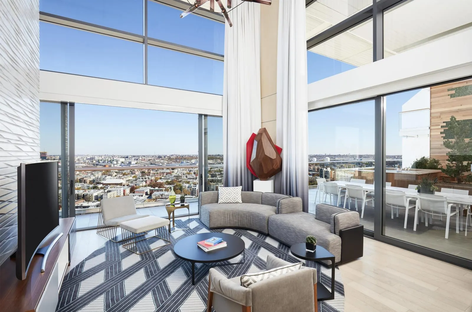 Vale Garden Residence at The William Vale: a double-height living room with two storeys of windows, a curved grey sofa, a round black coffee table, a faceted red and wood sculpture and a television, with a dining table and white chairs on the patio beyond the glass