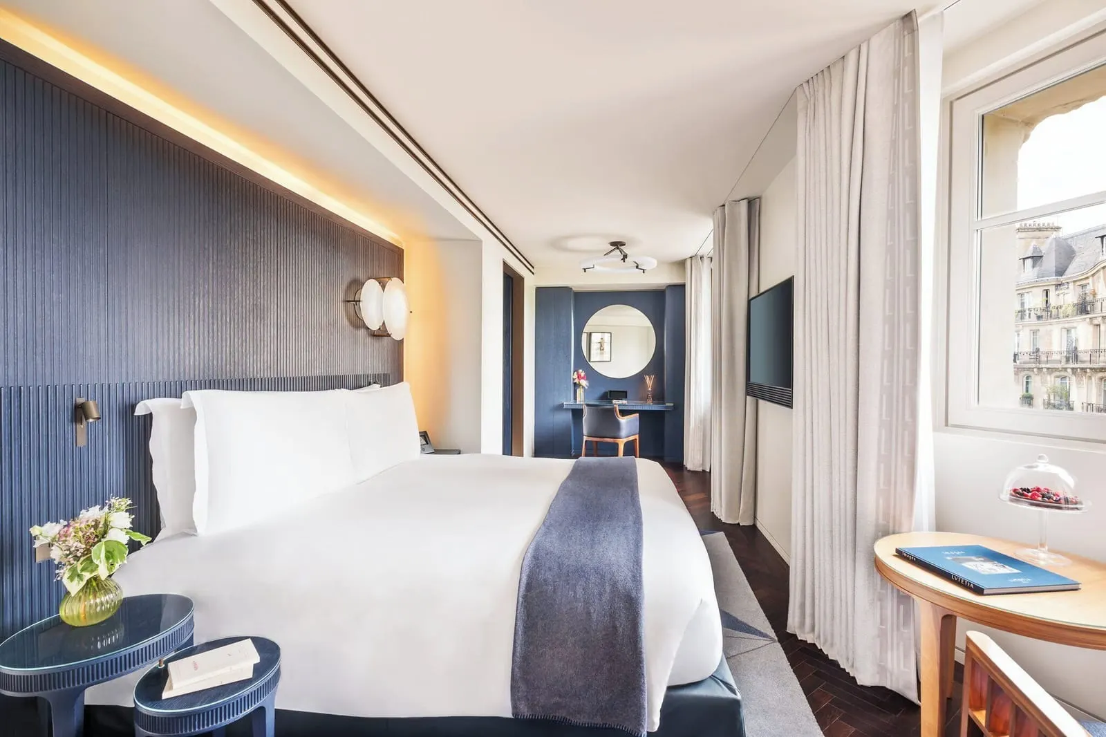 Family Suite at Mandarin Oriental Lutetia, Paris: a double bed against a dark blue fluted headboard, a dressing table with a round mirror, a wall-mounted television and a window over the street