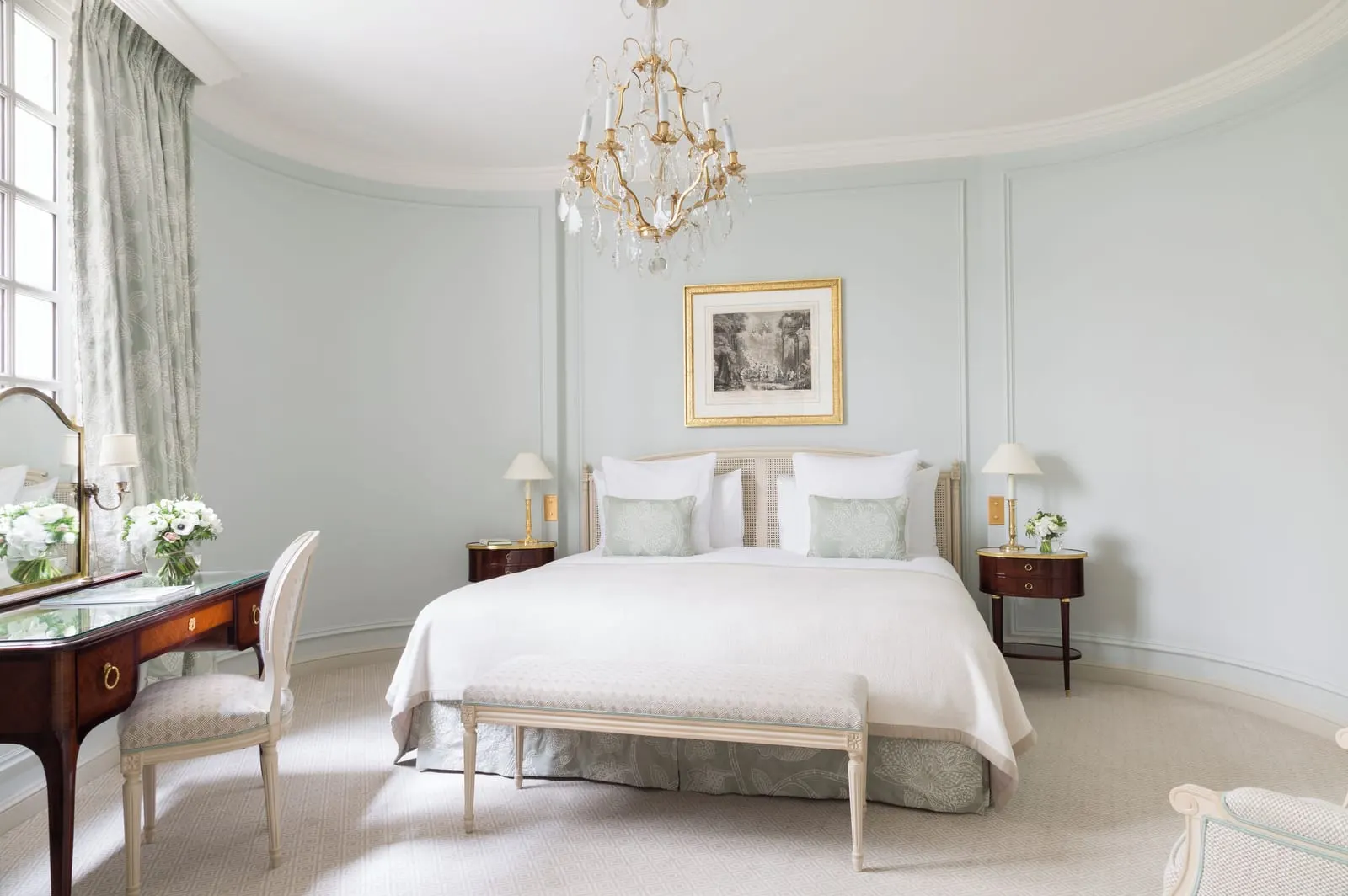 Deluxe Junior Suite at Le Bristol Paris: a bed against a curved, pale blue panelled wall under a crystal chandelier, with a writing desk by the window