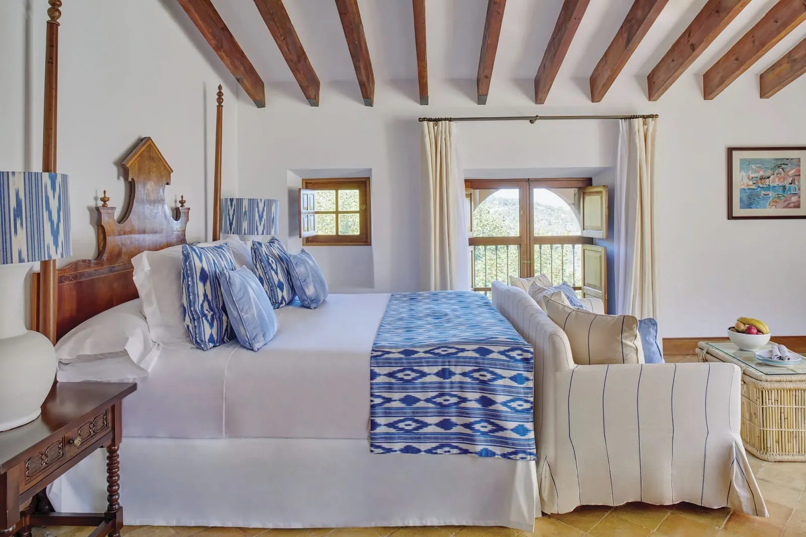 Superior Junior Suite at Belmond La Residencia: a bed with a carved wooden headboard and blue and white patterned cushions under wooden beams, with a striped sofa at its foot and an arched window with a balcony rail