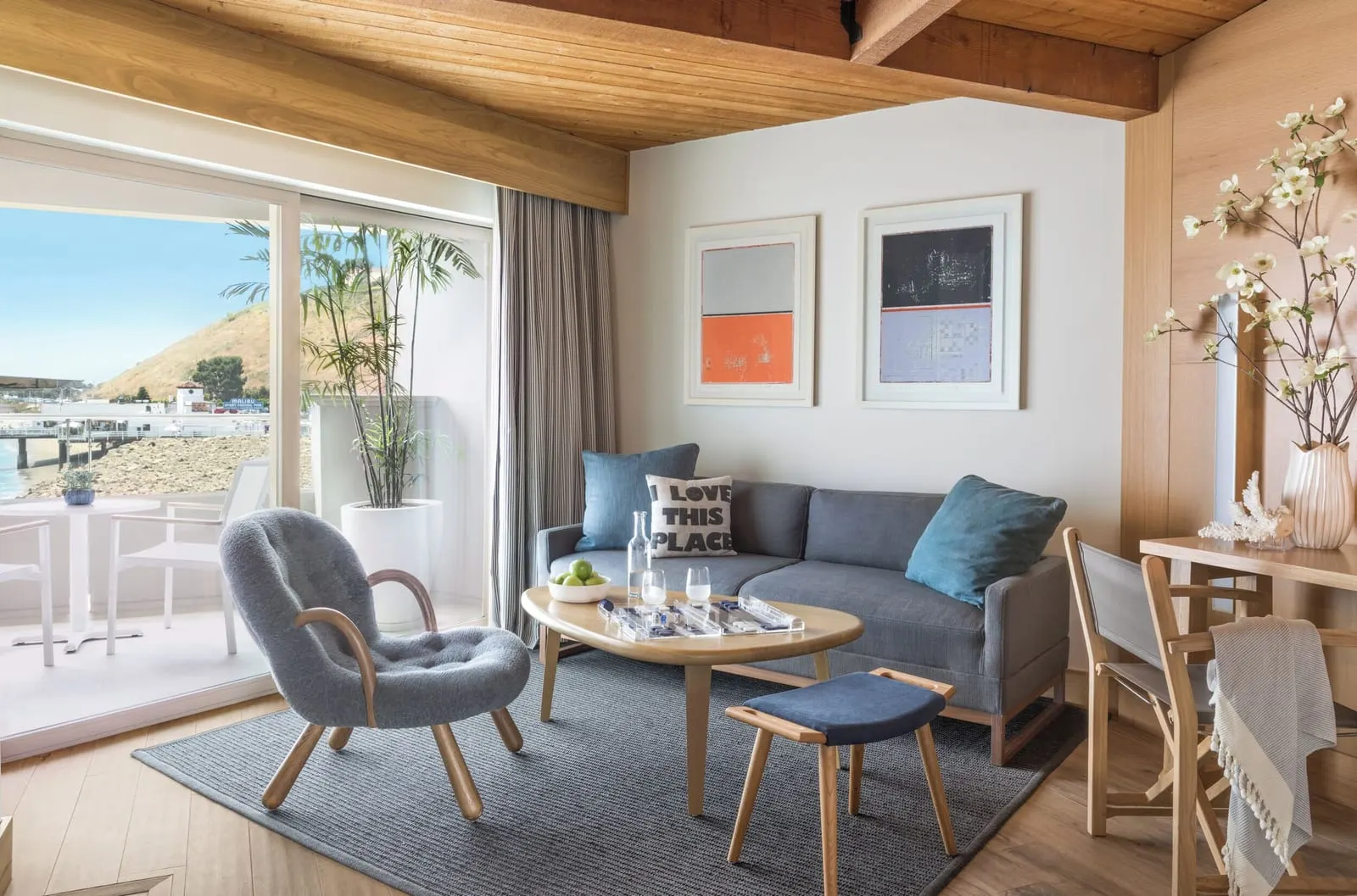 King Pier View Room at Malibu Beach Inn: a sofa, an armchair and a coffee table beside a sliding glass door onto a balcony, with the pier and a rocky shoreline beyond