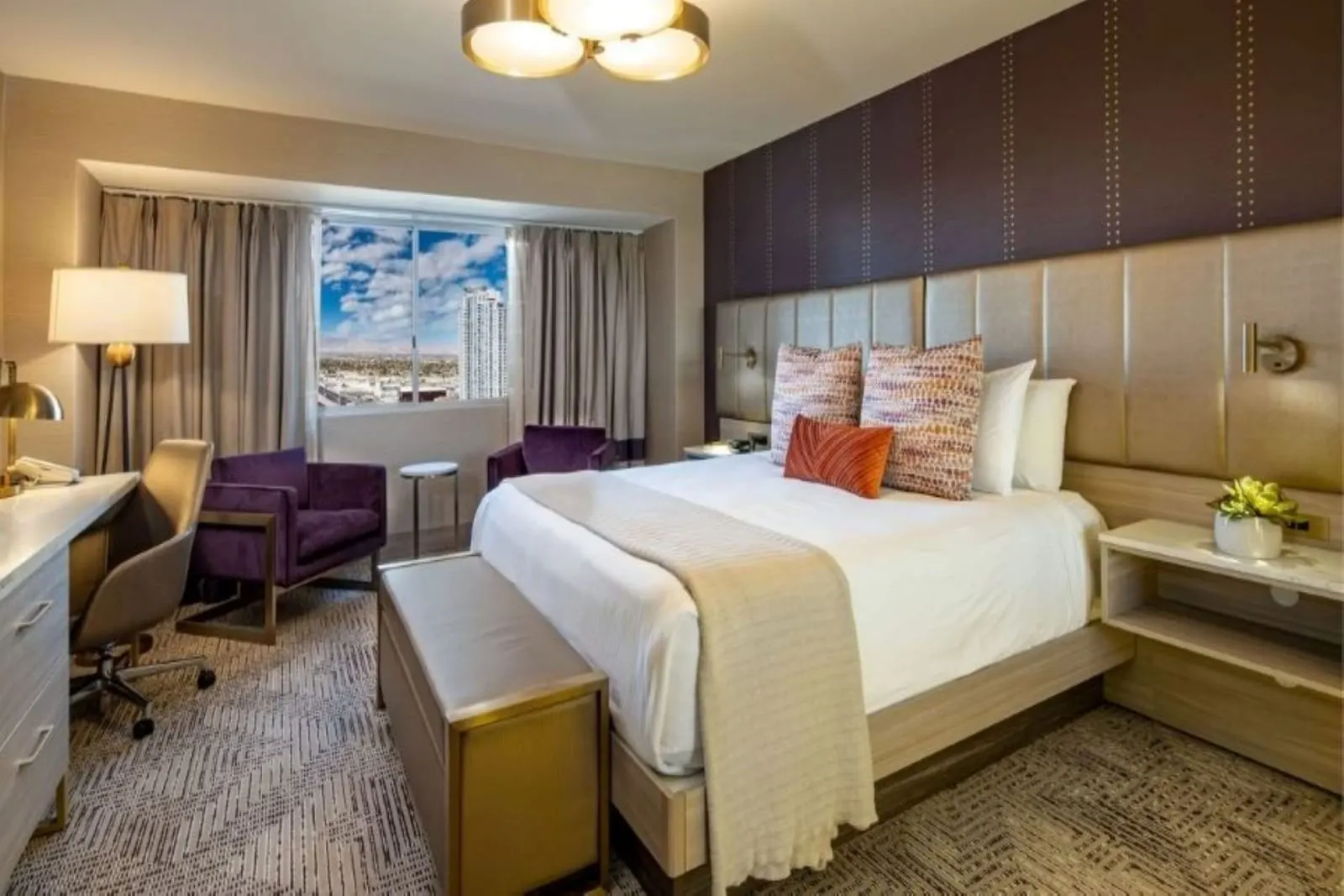 Marra Style King City View at SAHARA Las Vegas: a king bed with a padded headboard and a bronze bench, two purple armchairs, a desk and a window onto the city