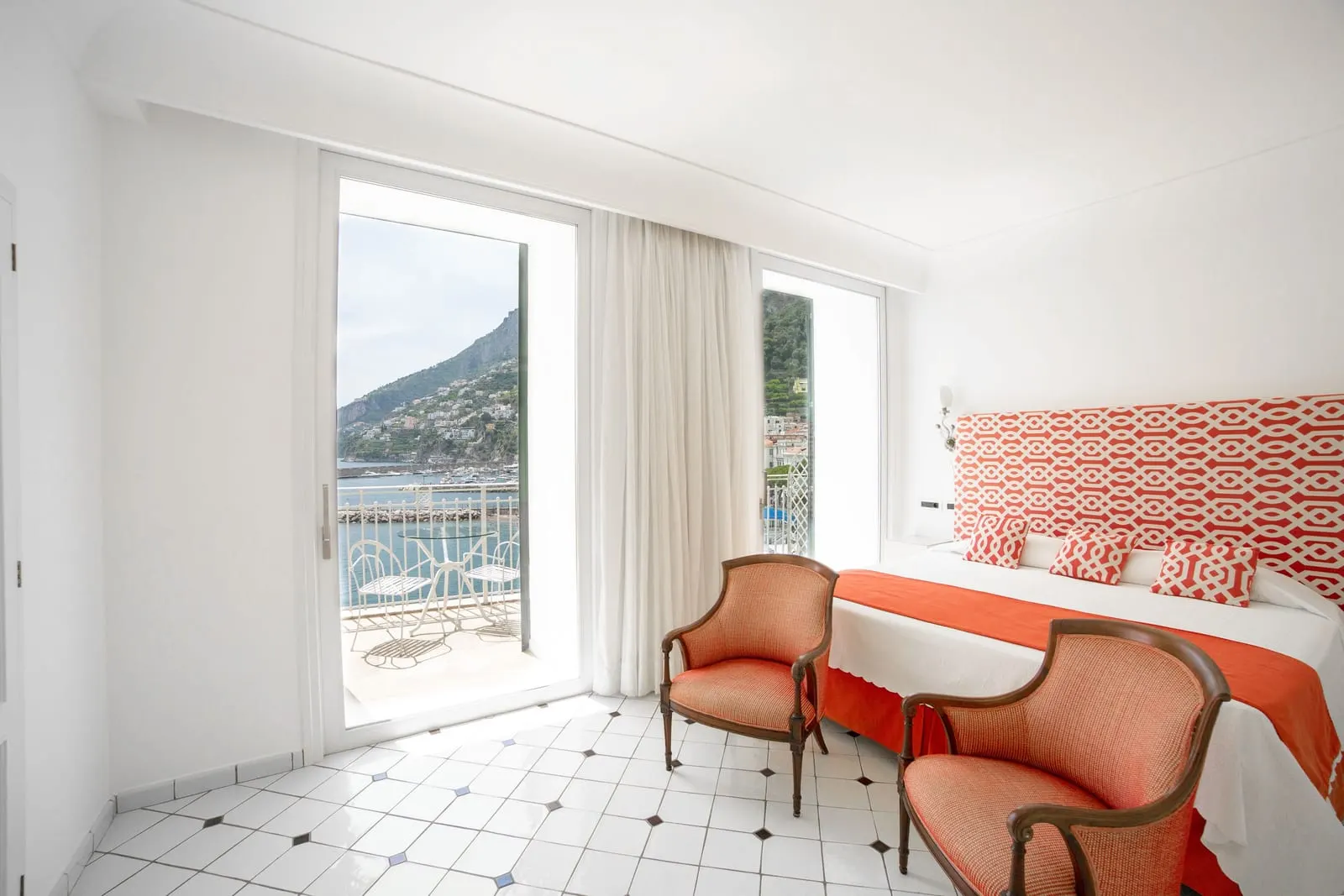 Junior Suite（Hotel Marina Riviera）：ダブルベッドとアームチェア2脚が、湾を見渡すテーブルと椅子付きのバルコニーへのガラス扉の脇にある