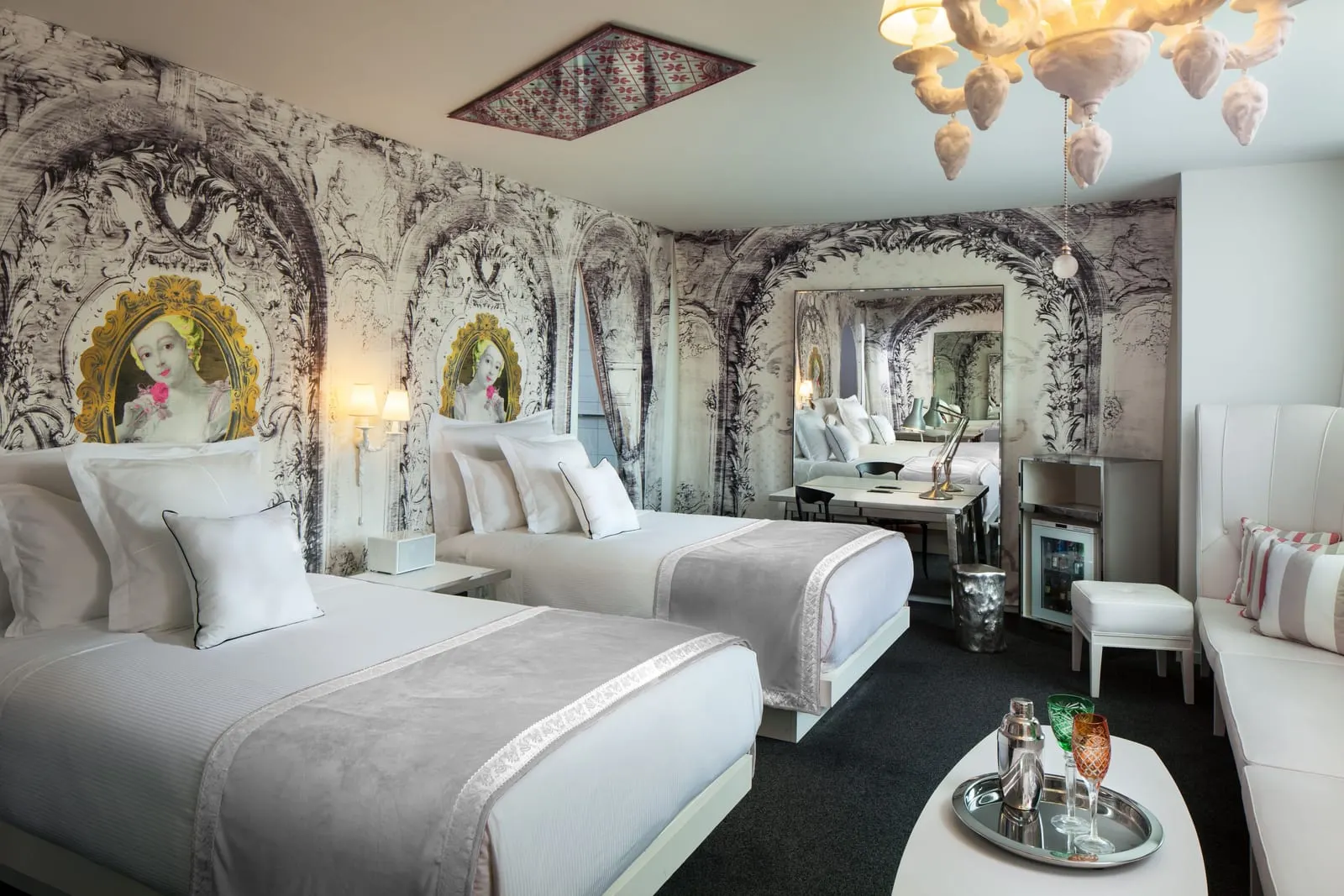 Alexandria Iconic Queens at SAHARA Las Vegas: two queen beds against a black and white baroque-print wall with framed portraits, a desk in front of a tall mirror, a mini fridge and a white banquette with a low table