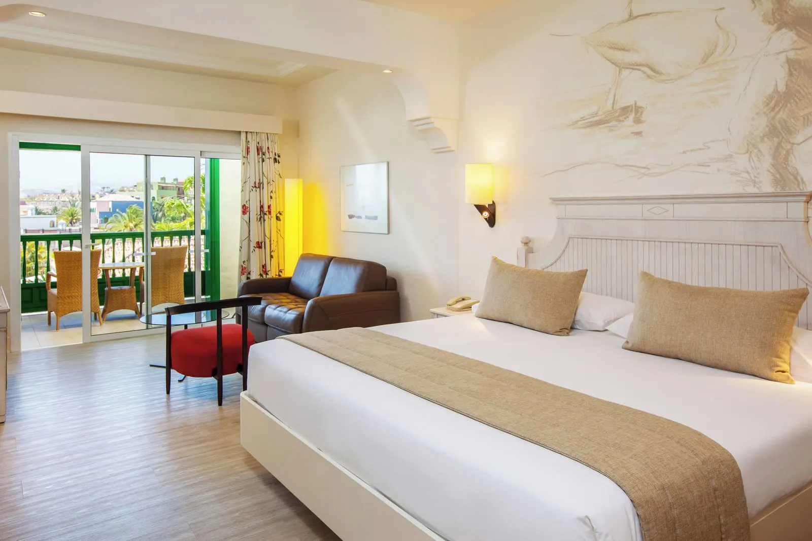 Double Standard View at Lopesan Villa del Conde Resort & Thalasso: a king bed under a sketched mural, a small leather sofa and a red chair, and sliding doors to a balcony with two wicker chairs and a table