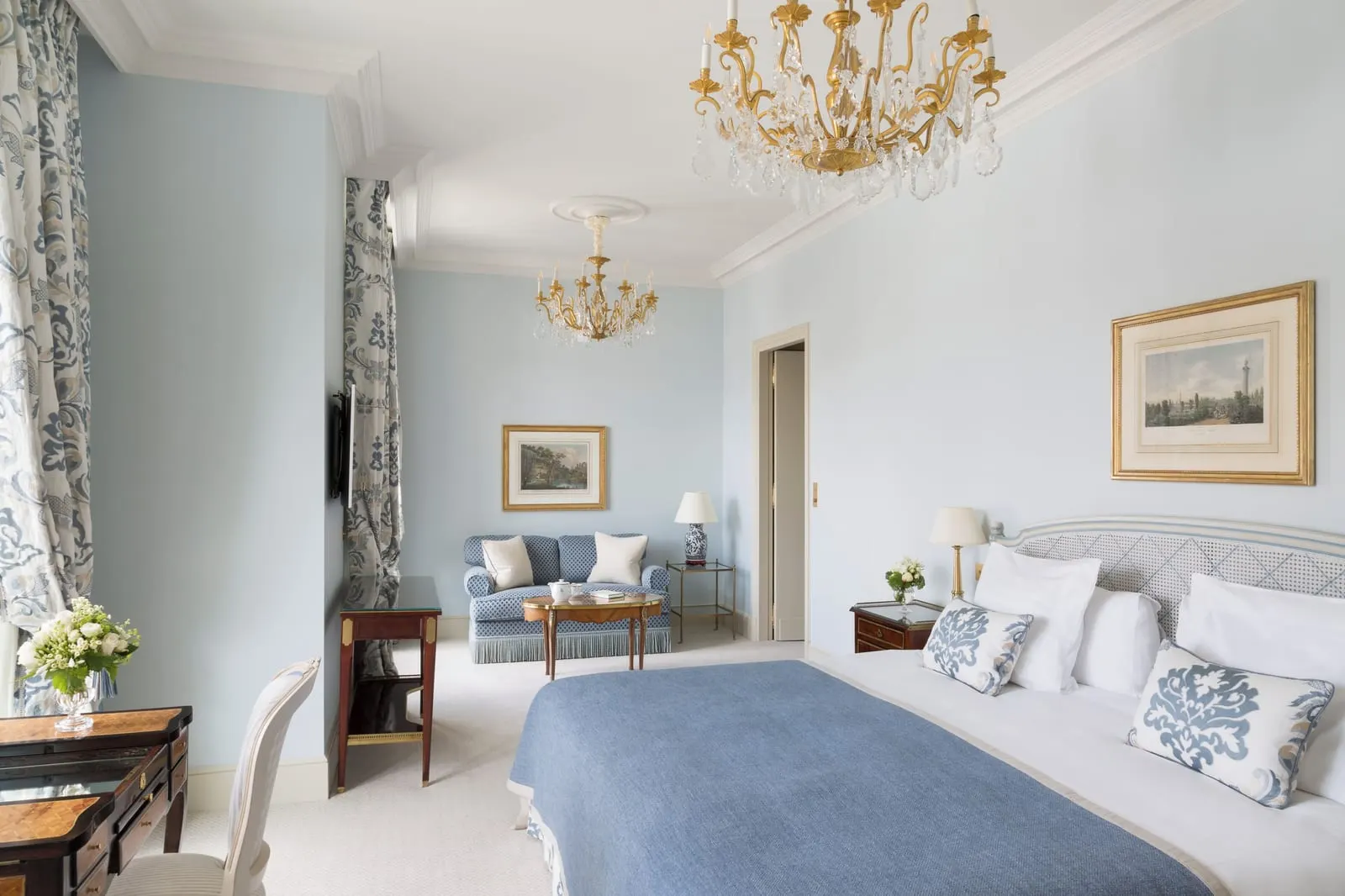 Junior Suite with Skyline view at Le Bristol Paris: a bed with a blue throw in the foreground and a sitting area with a blue sofa beyond, under two crystal chandeliers