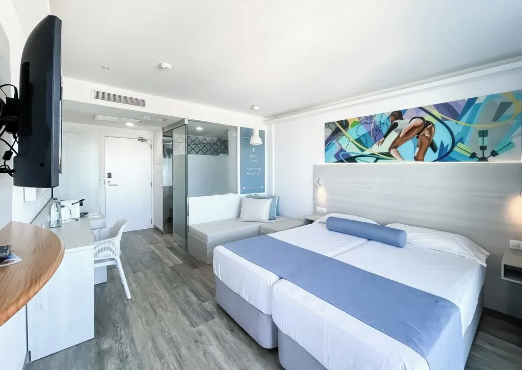 Sky Sea View at Corralejo Surfing Colors Hotel & Apartments: two single beds made up as one under a colourful mural, with a desk, a wall-mounted television and a glass-walled bathroom by the door.