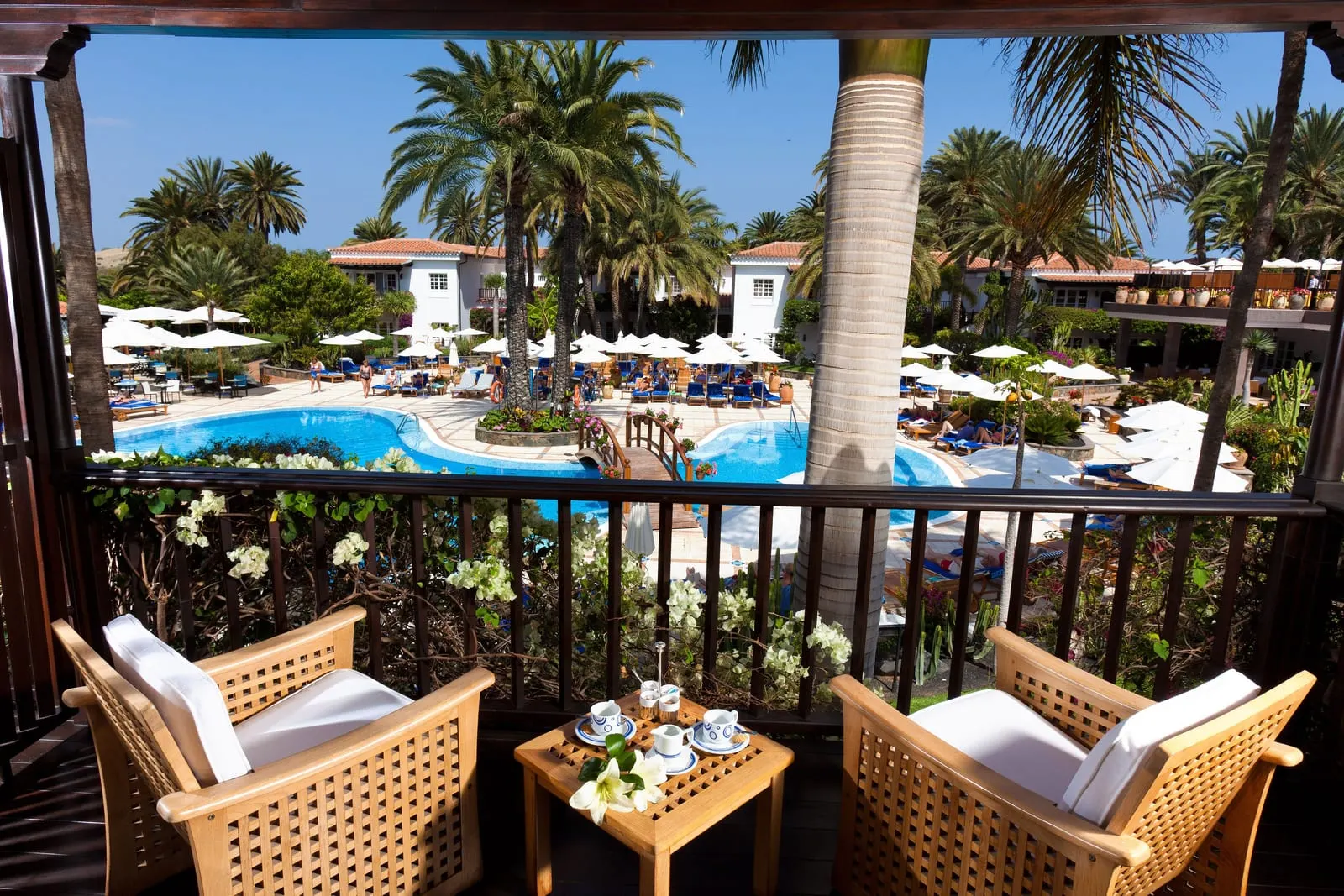 Deluxe Room at Seaside Grand Hotel Residencia: a balcony with two teak armchairs and a coffee table, looking through palms to the pool